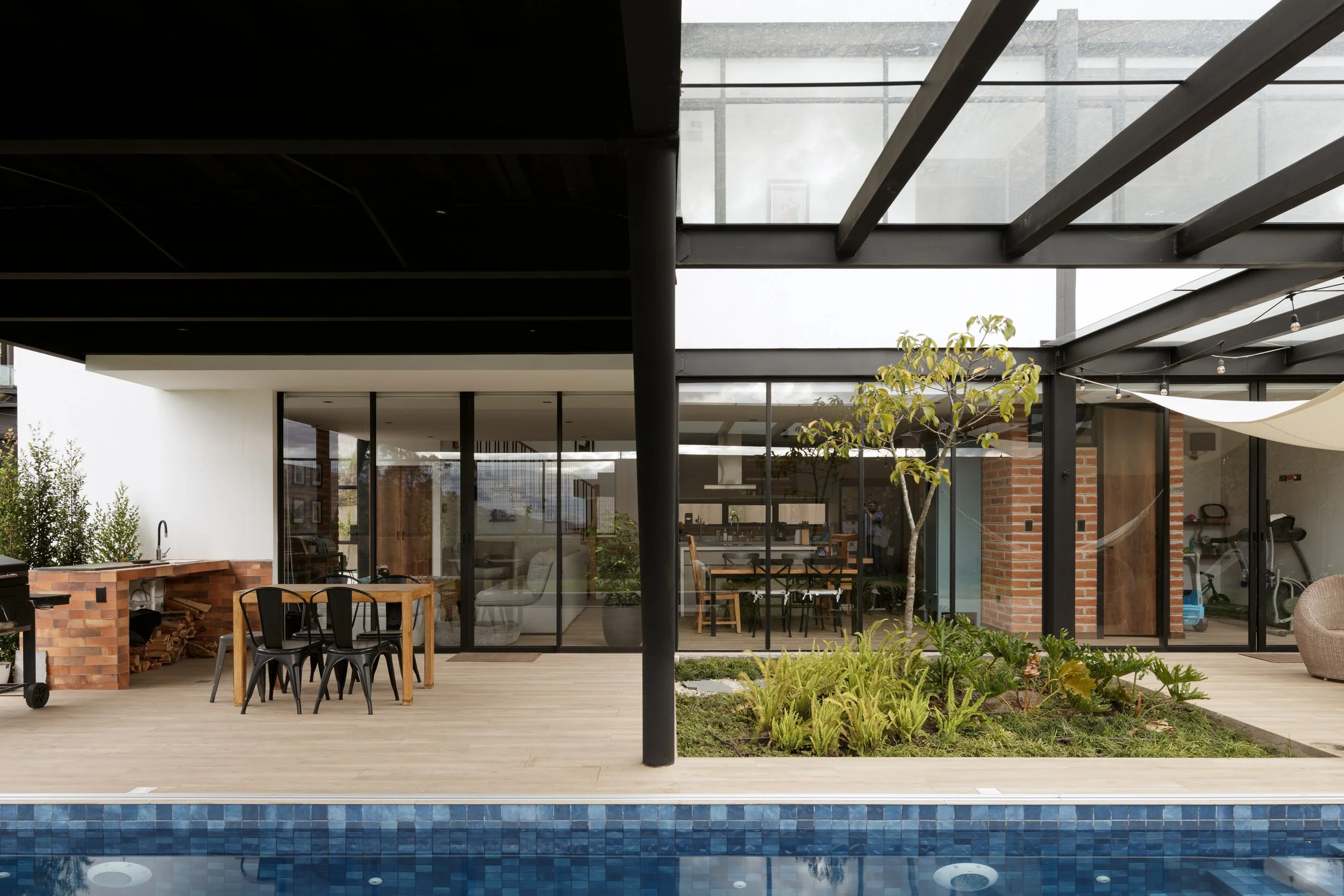 Área de patio exterior con piscina y jardín, estructura moderna con paredes de vidrio y techo de metal, vista al interior con muebles de comedor y árbol pequeño.