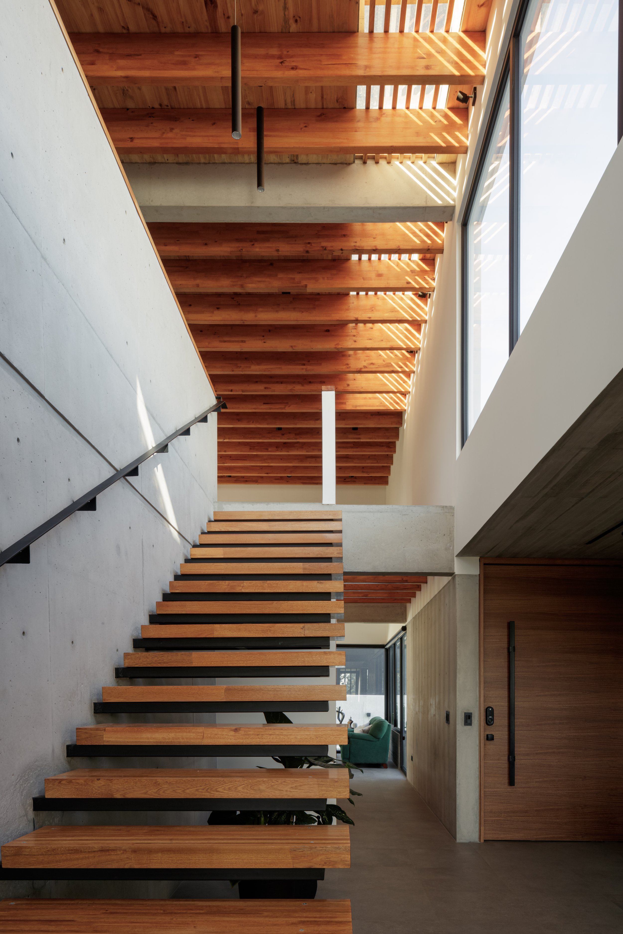 Interior moderno de una casa con escalera de madera y bloques de concreto, ventanas grandes que dejan entrar luz natural, y detalles en madera y cemento.