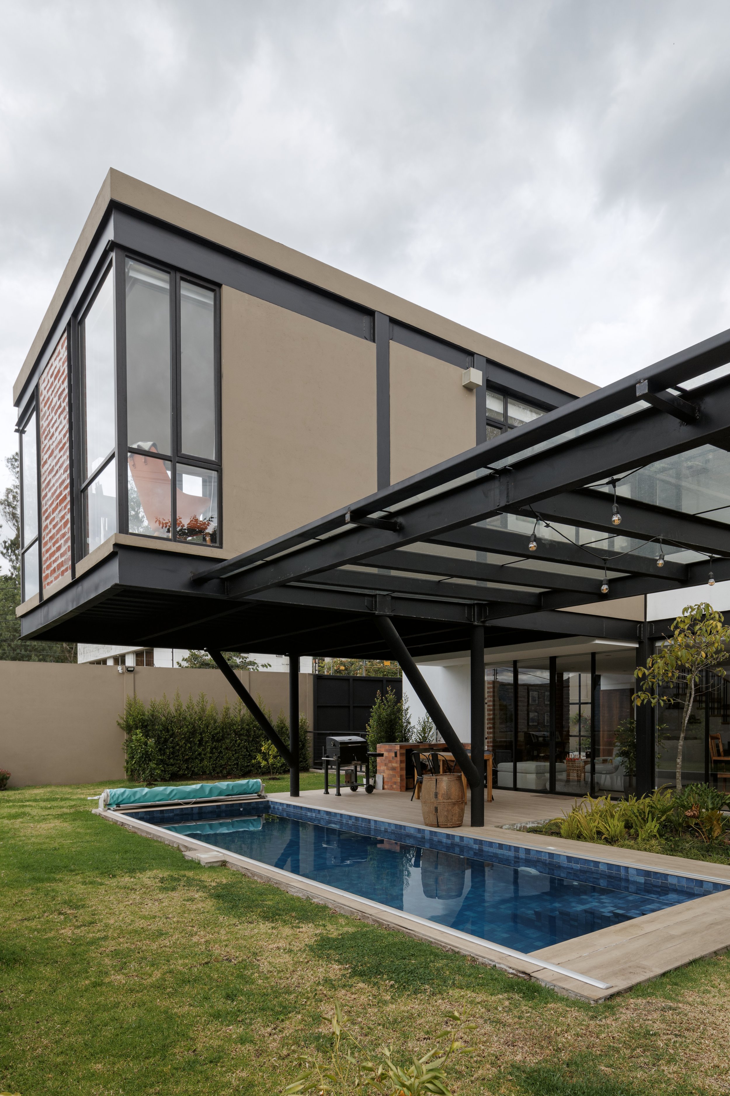Casa moderna de varios pisos con grandes ventanales, jardín con alberca, mobiliario de exterior, parrilla y árboles.