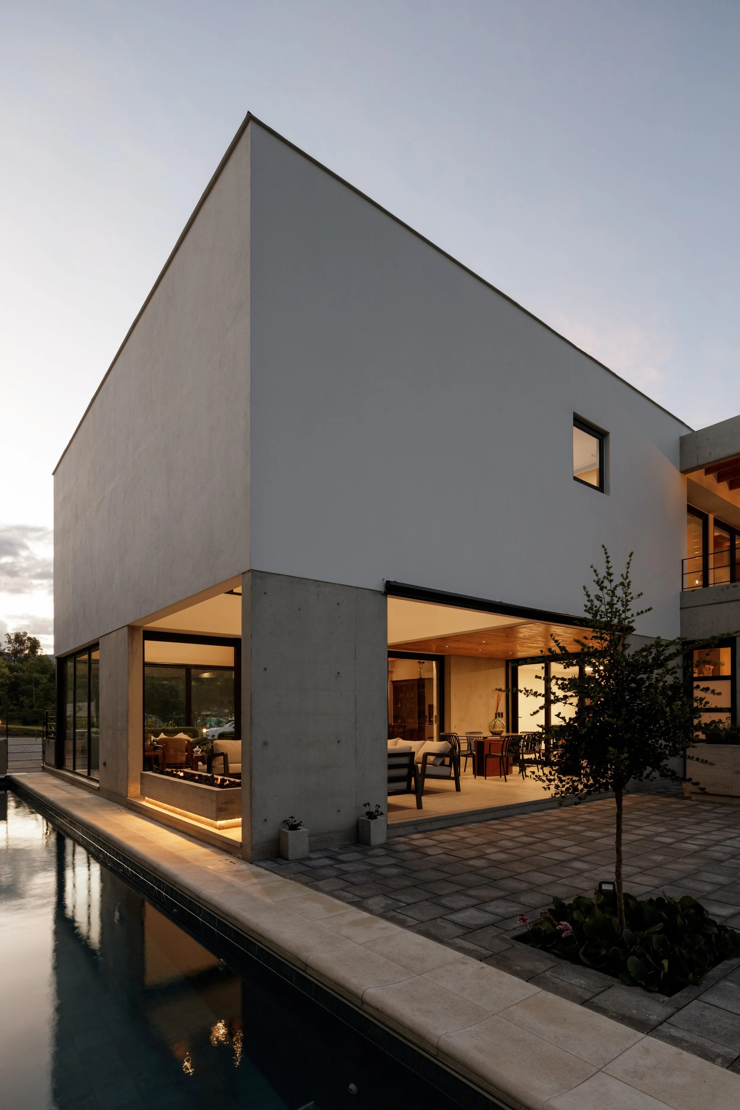Casa moderna de varios pisos con amplias ventanas, patio y piscina, ubicada en un entorno con árboles, durante el atardecer con luz cálida y cielo despejado.