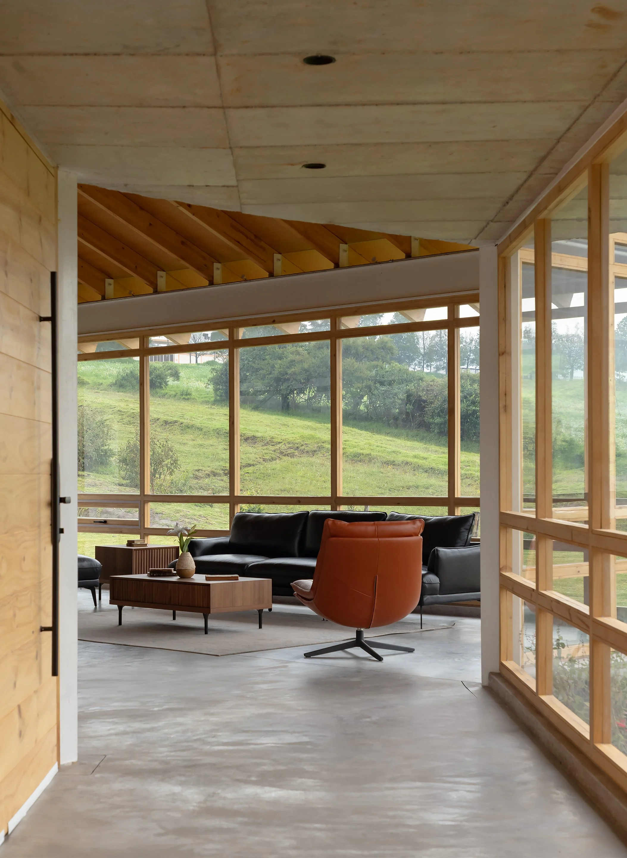 Sala de estar moderna con grandes ventanales, sillones negros y una silla naranja, con vista al campo y árboles verdes.