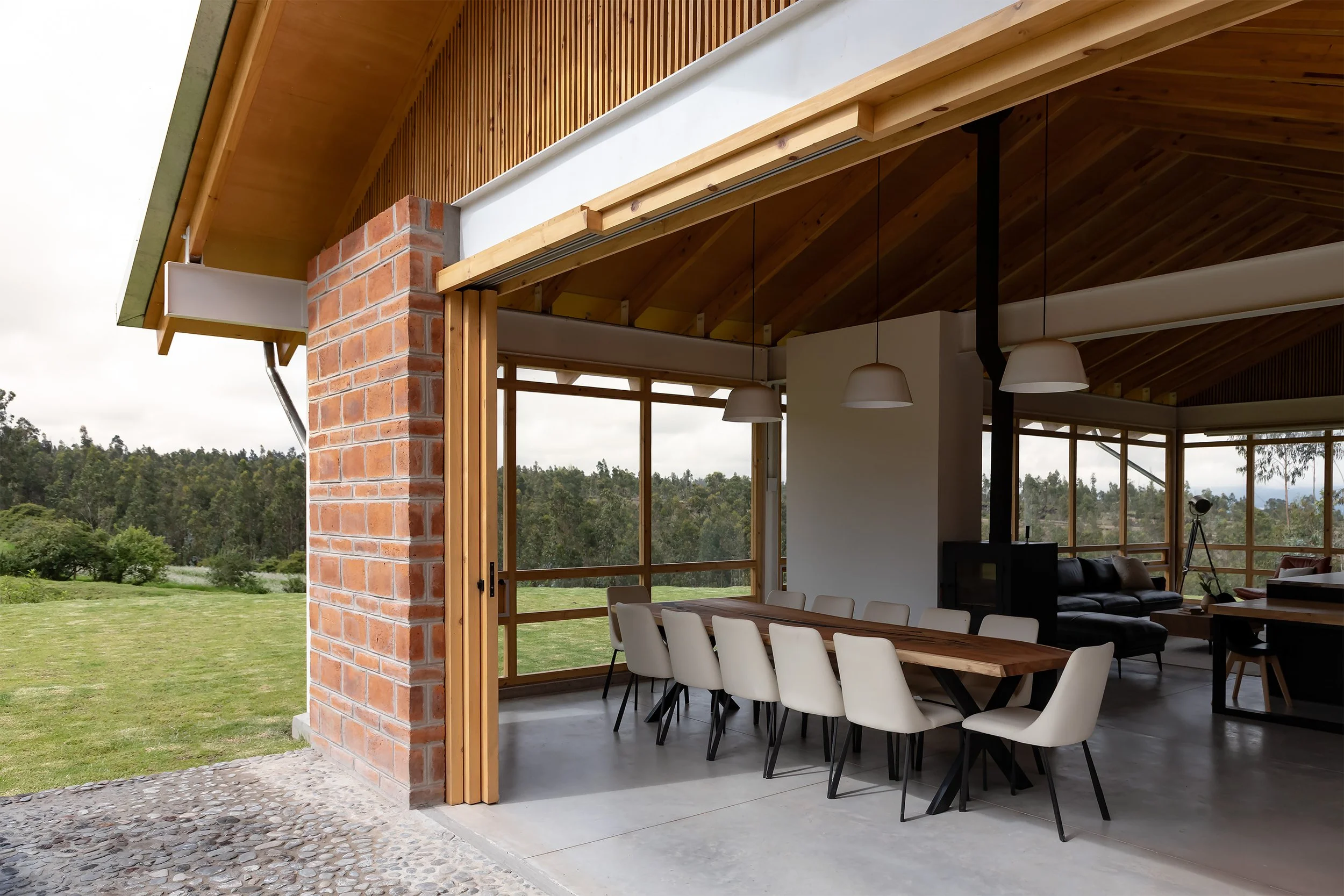 Interiores de una casa moderna con paredes de vidrio grandes, techo de madera, y muebles de comedor con sillas blancas, con vista a un área verde exterior.