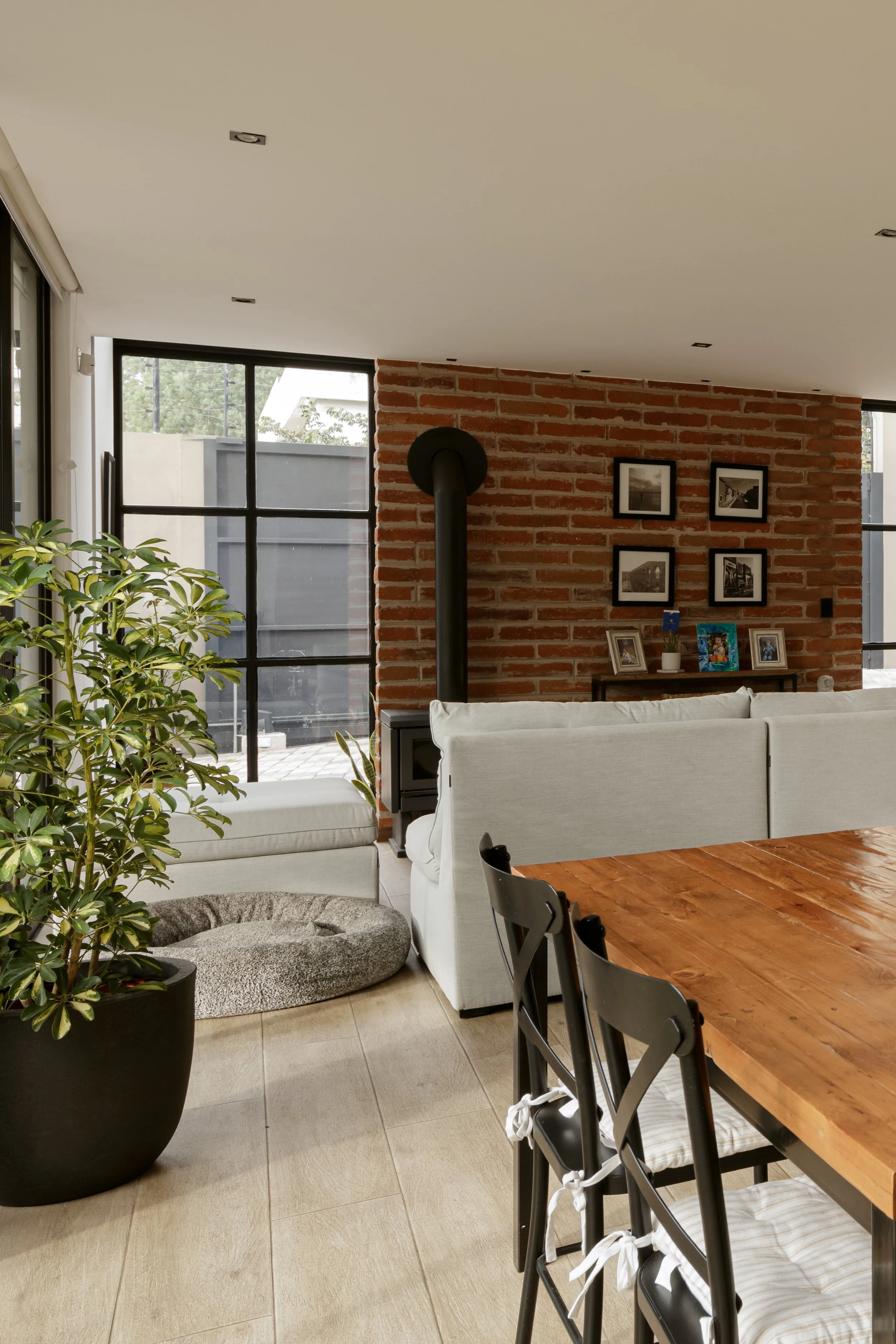Sala moderna con sofá blanco, mesa de madera y paredes de ladrillo con cuadros, ventanas grandes y plantas decorativas.