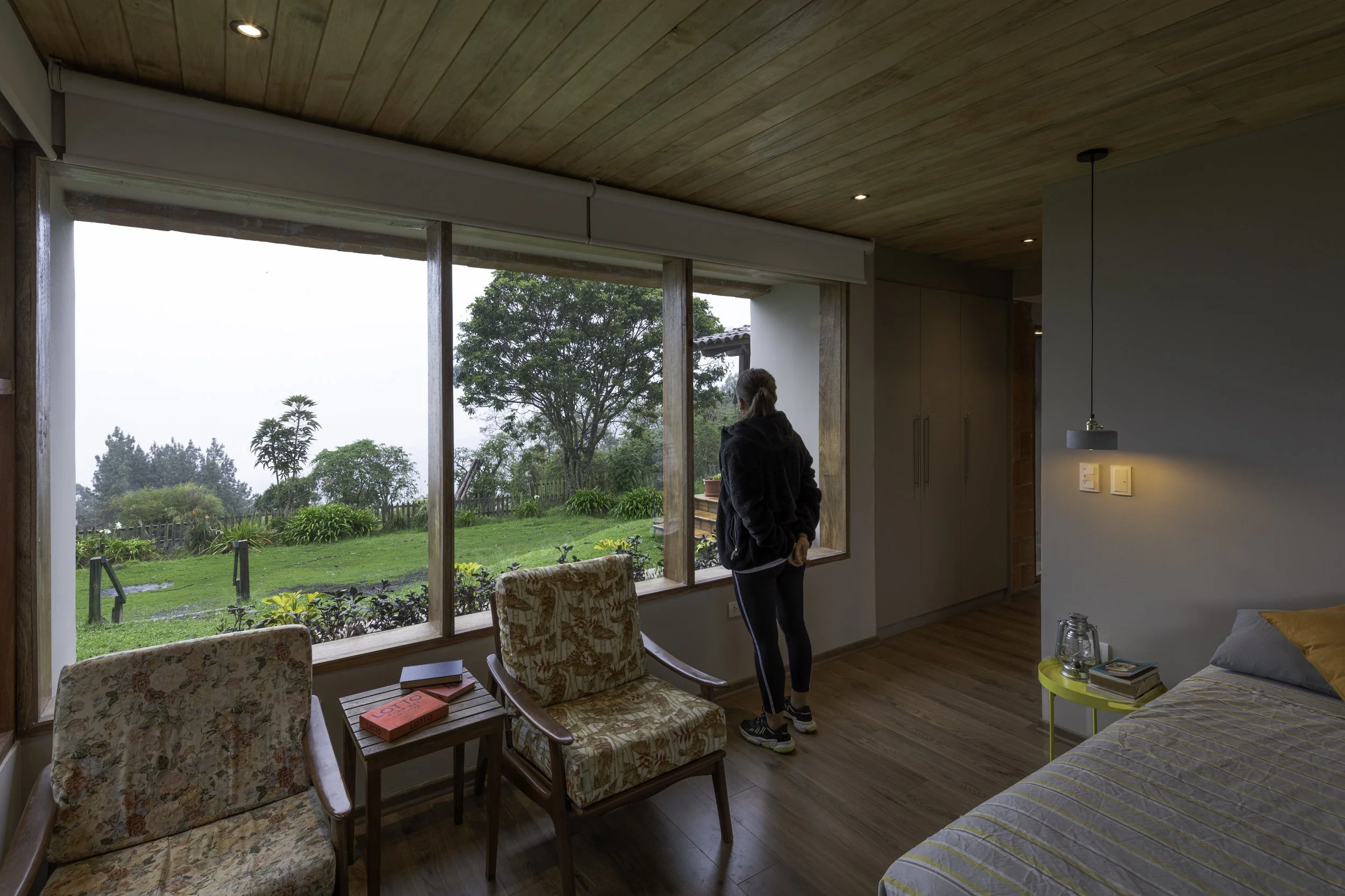 Una mujer de espaldas, de pie, mirando por una ventana grande que muestra un jardín con árboles y plantas verdes, en una habitación con techo de madera y paredes blancas. Hay dos sillas con tapizado floral y mesas pequeñas dentro de la habitación.