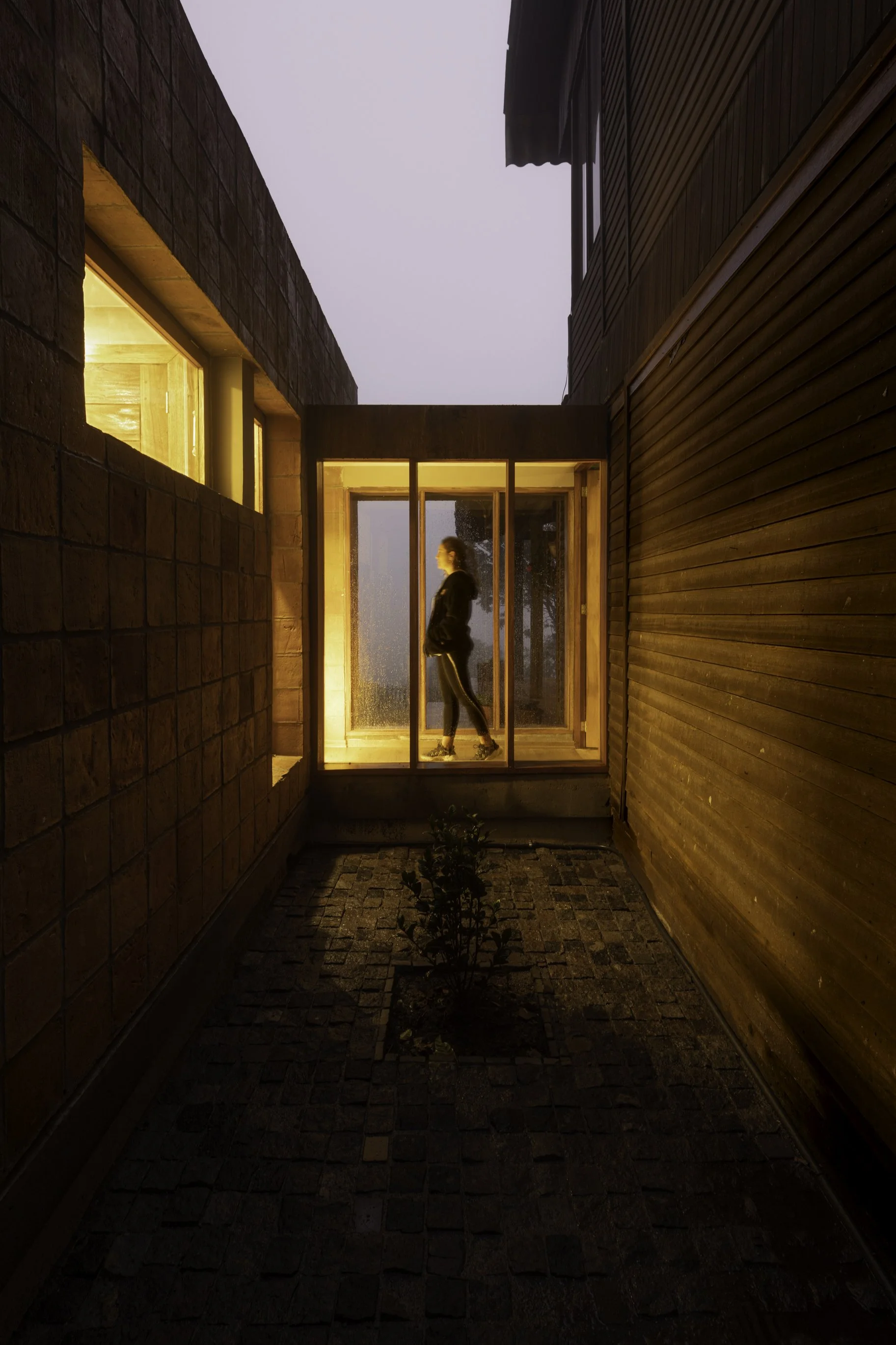 Pasillo exterior con muros de ladrillo y madera, con iluminación cálida en el interior y una persona caminando en la puerta de cristal.