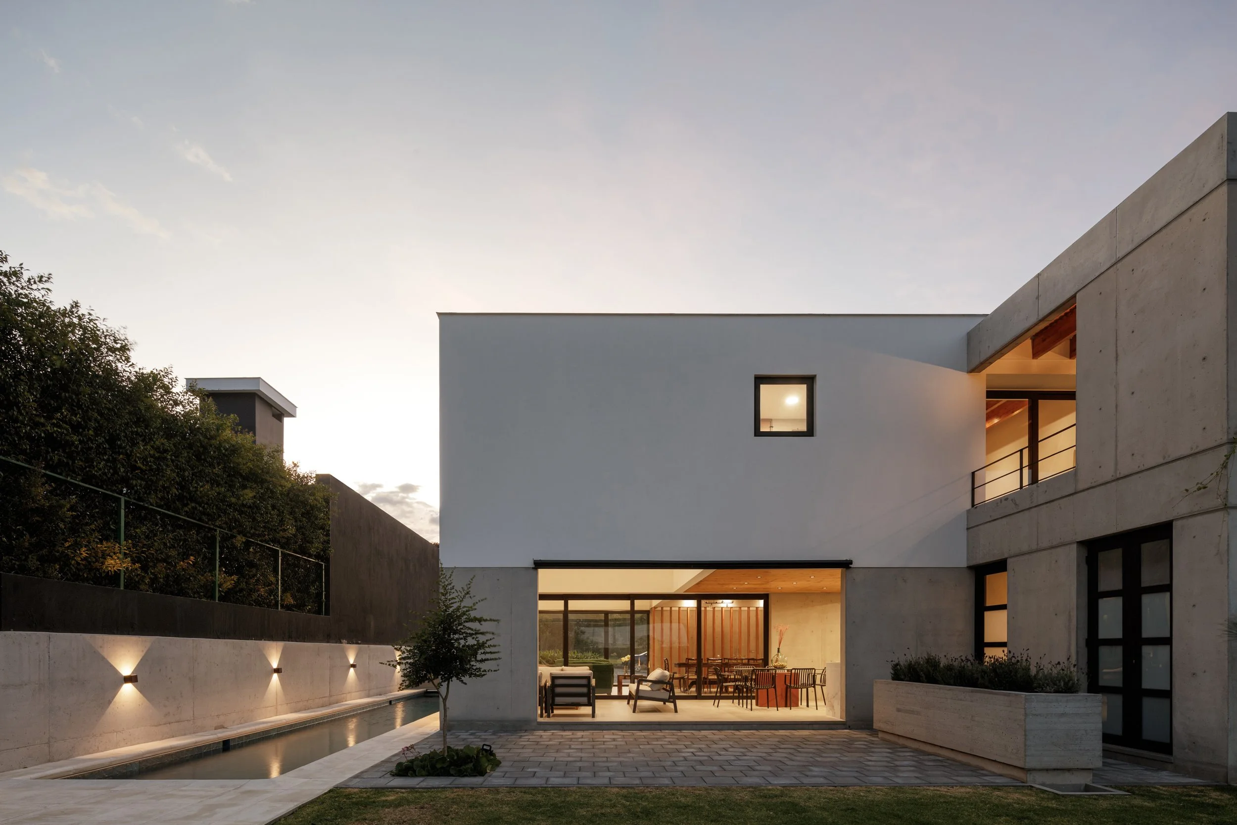 Fachada moderna de casa con muro de concreto, ventanas grandes y área de terraza con muebles, junto a piscina y árboles, al atardecer