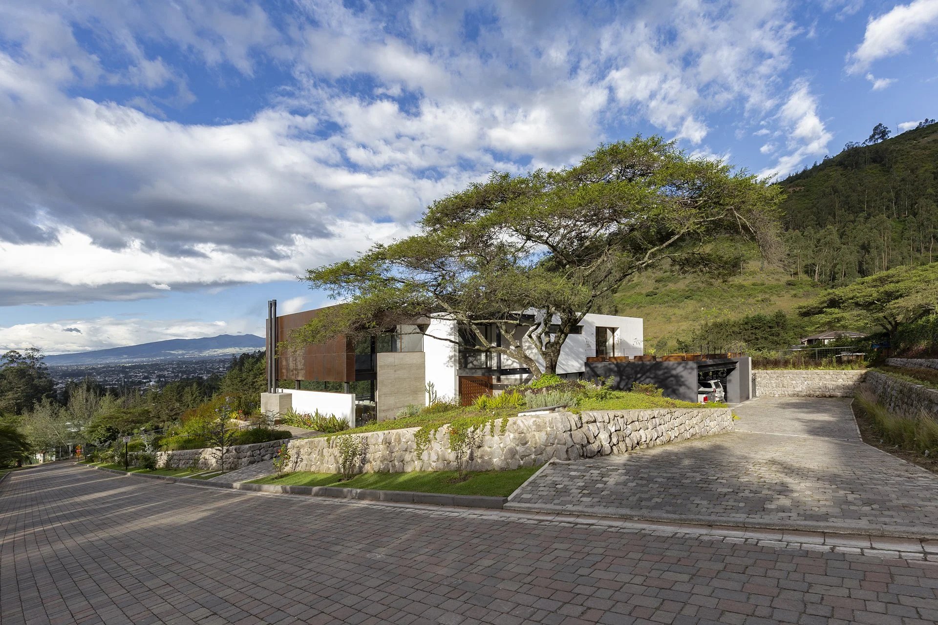 Casa moderna con árboles y colinas verdes de fondo, caminos de piedra y autos en el garaje.