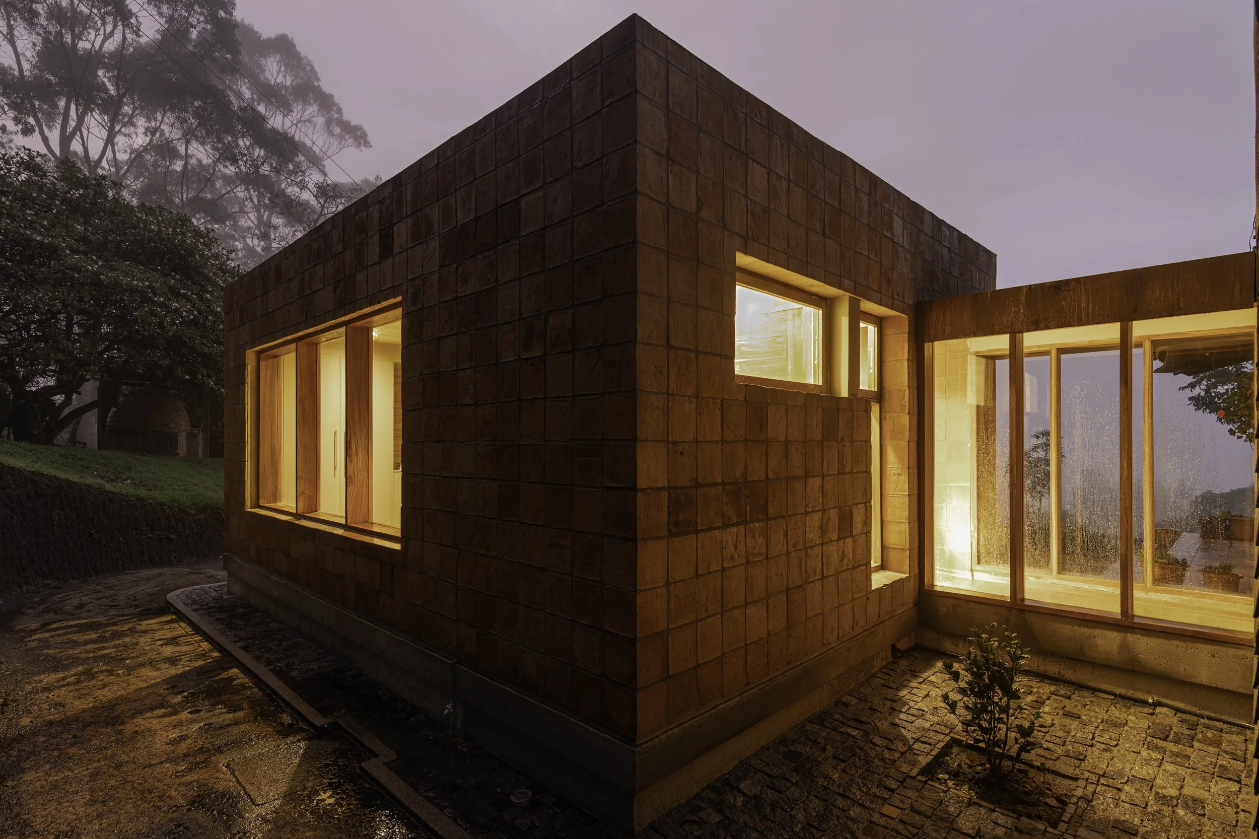 Una casa moderna construida con bloques de ladrillo, con ventanas iluminadas y rodeada de árboles en un ambiente nublado al atardecer.