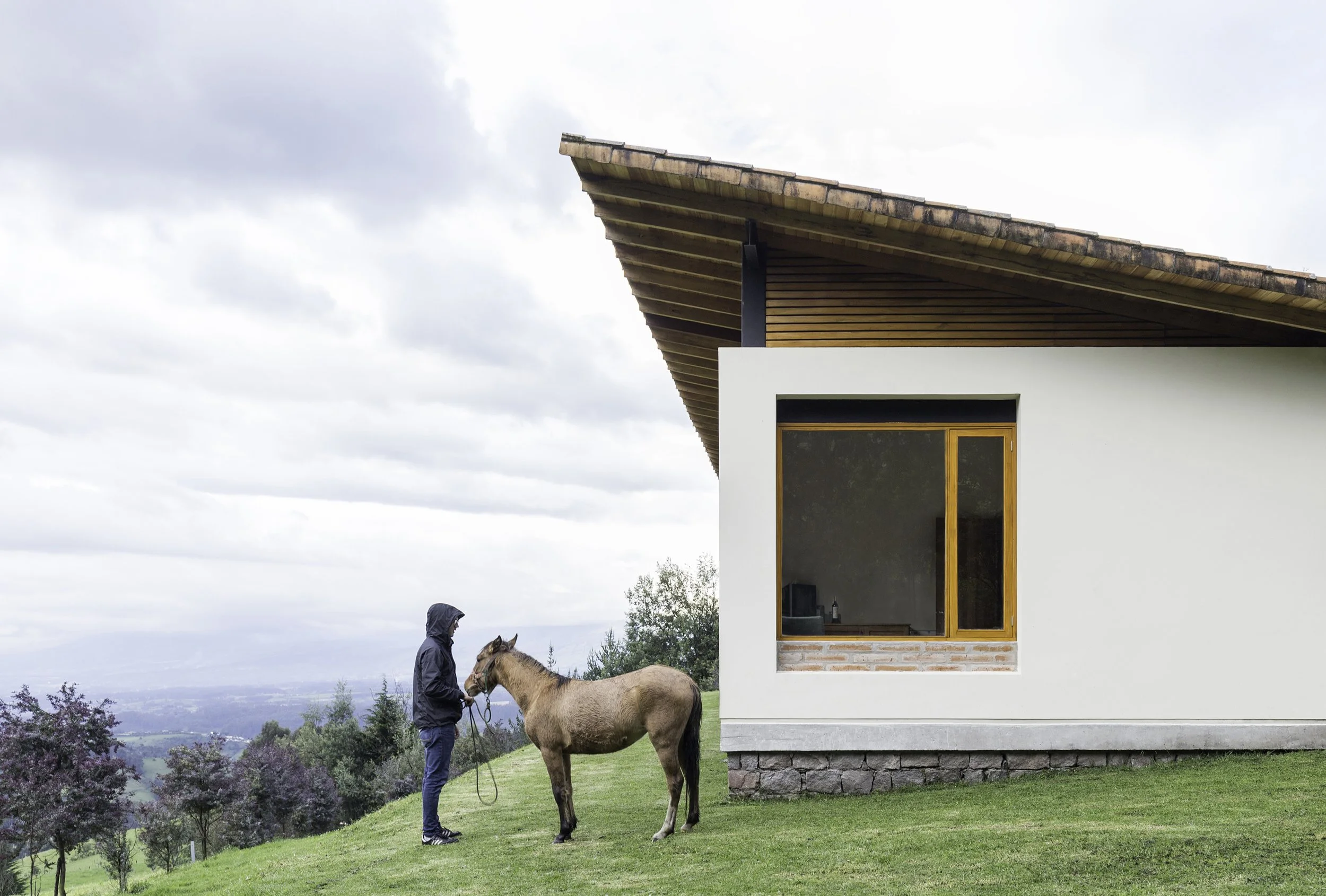Hombre con chaqueta de lluvia sostiene las riendas de un caballo en un césped verde frente a una casa moderna con paredes blancas, ventanas de madera y un techo inclinado de tejas, en un paisaje montañoso con árboles y cielos nublados.