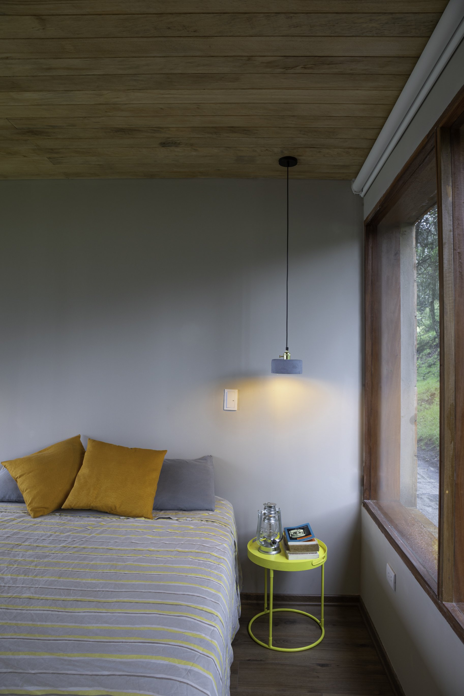 Habitación con paredes blancas y techo de madera, cama con sábanas a rayas en tonos gris y amarillo, con tres almohadas en tonos mostaza y gris, a la derecha una ventana grande con marco de madera, y una lámpara de mesa amarilla con libros y una lámp