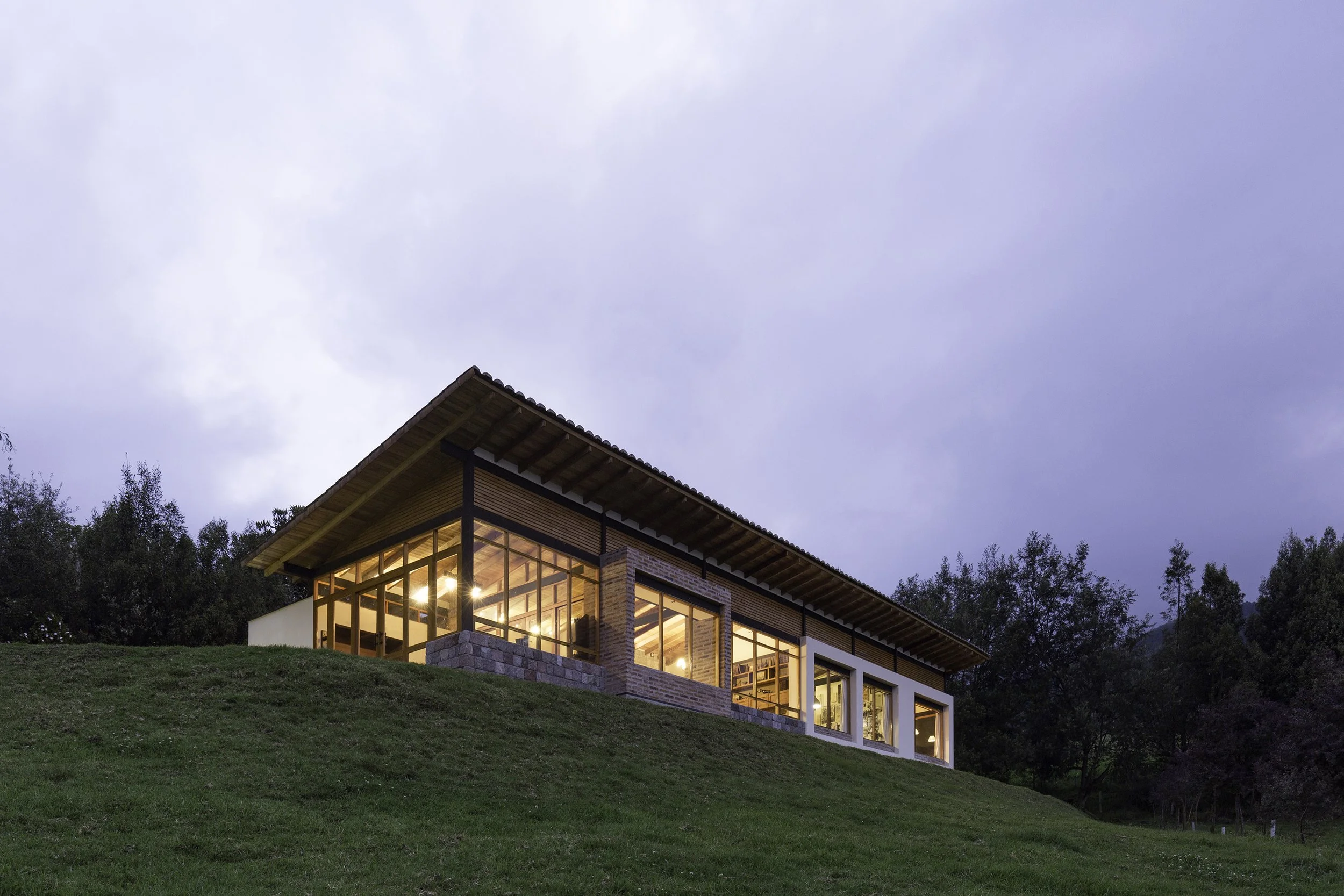 Casa moderna de madera y ladrillo con grandes ventanas iluminadas, ubicada en un terreno con césped y rodeada de árboles, vista nocturna con cielo nublado.