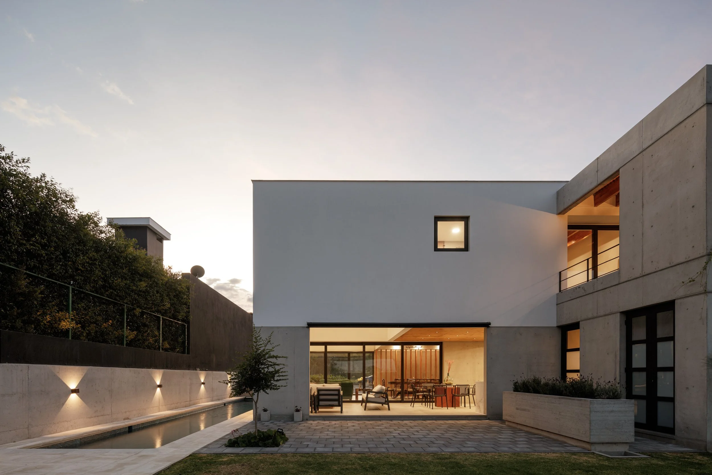 Casa moderna con grande ventana y patio trasero iluminado por luces, piscina y muebles de exterior, en hora del atardecer.