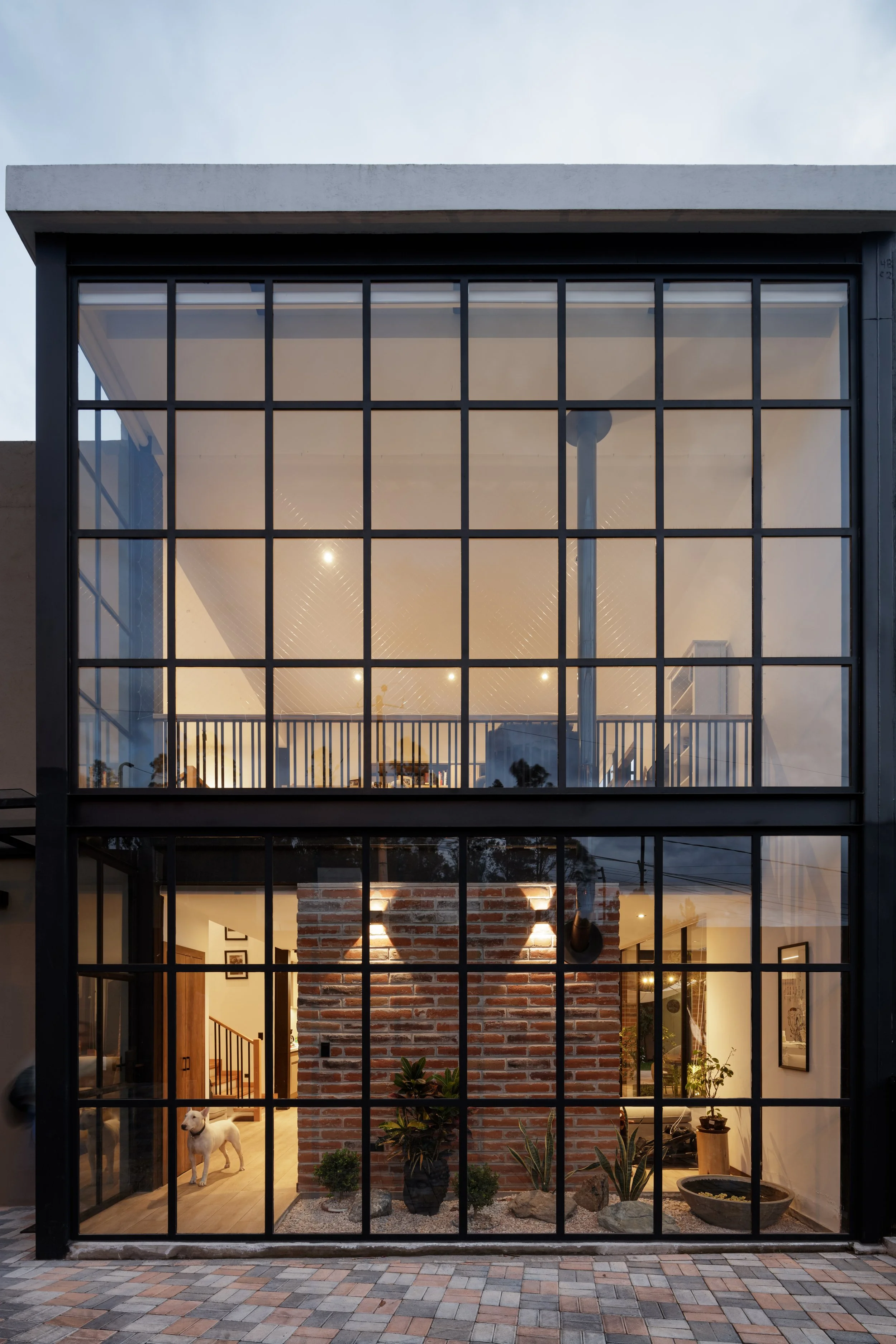 Vista frontal de una casa moderna con estructura de acero y grandes ventanas de vidrio, interior con paredes de ladrillo expuesto, plantas y decoración, y perros en la entrada.