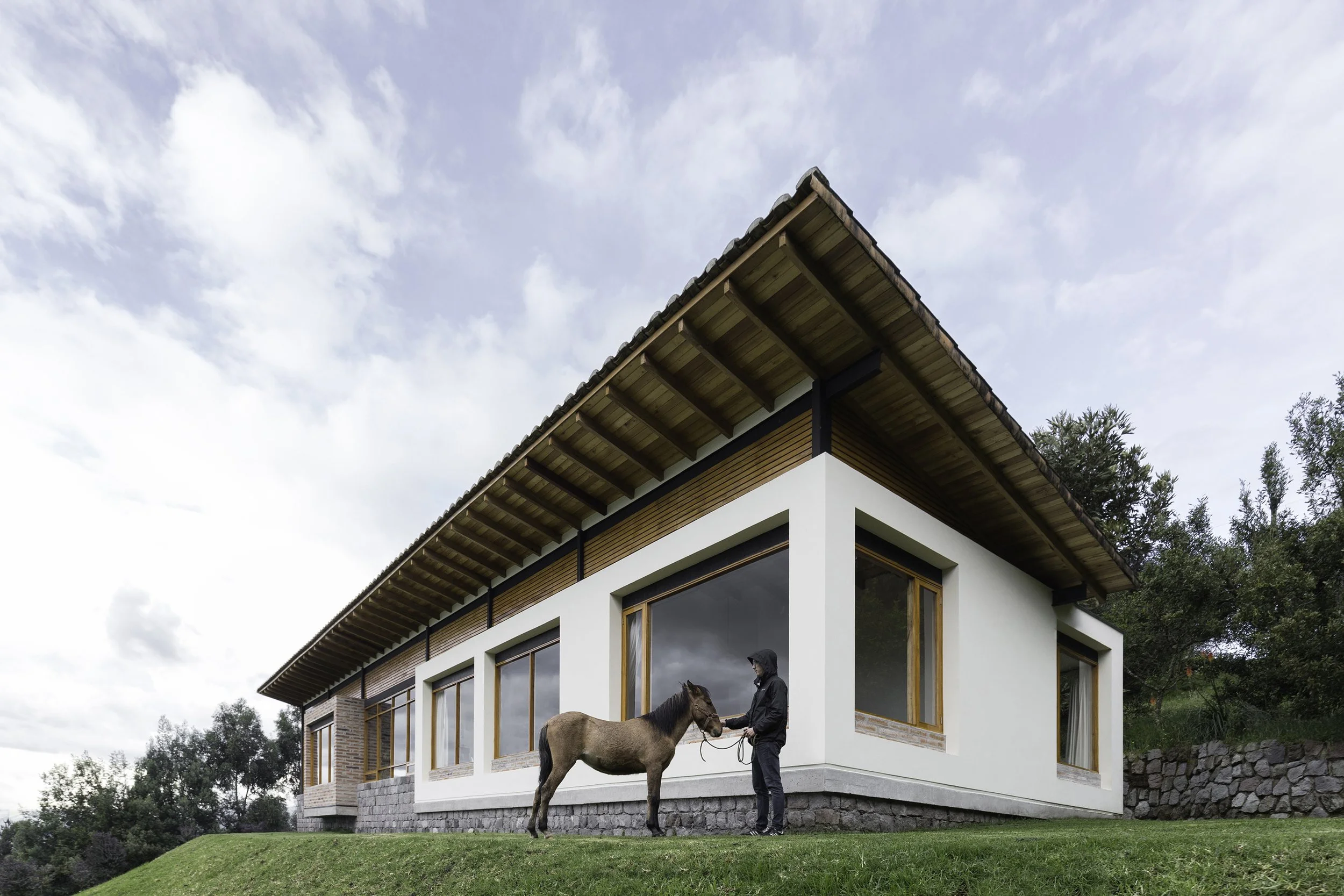 Casa moderna de dos pisos con grandes ventanas de vidrio y techo de tejas de cerámica, rodeada de áreas verdes, en un día nublado. Un hombre con un caballo en la entrada. 