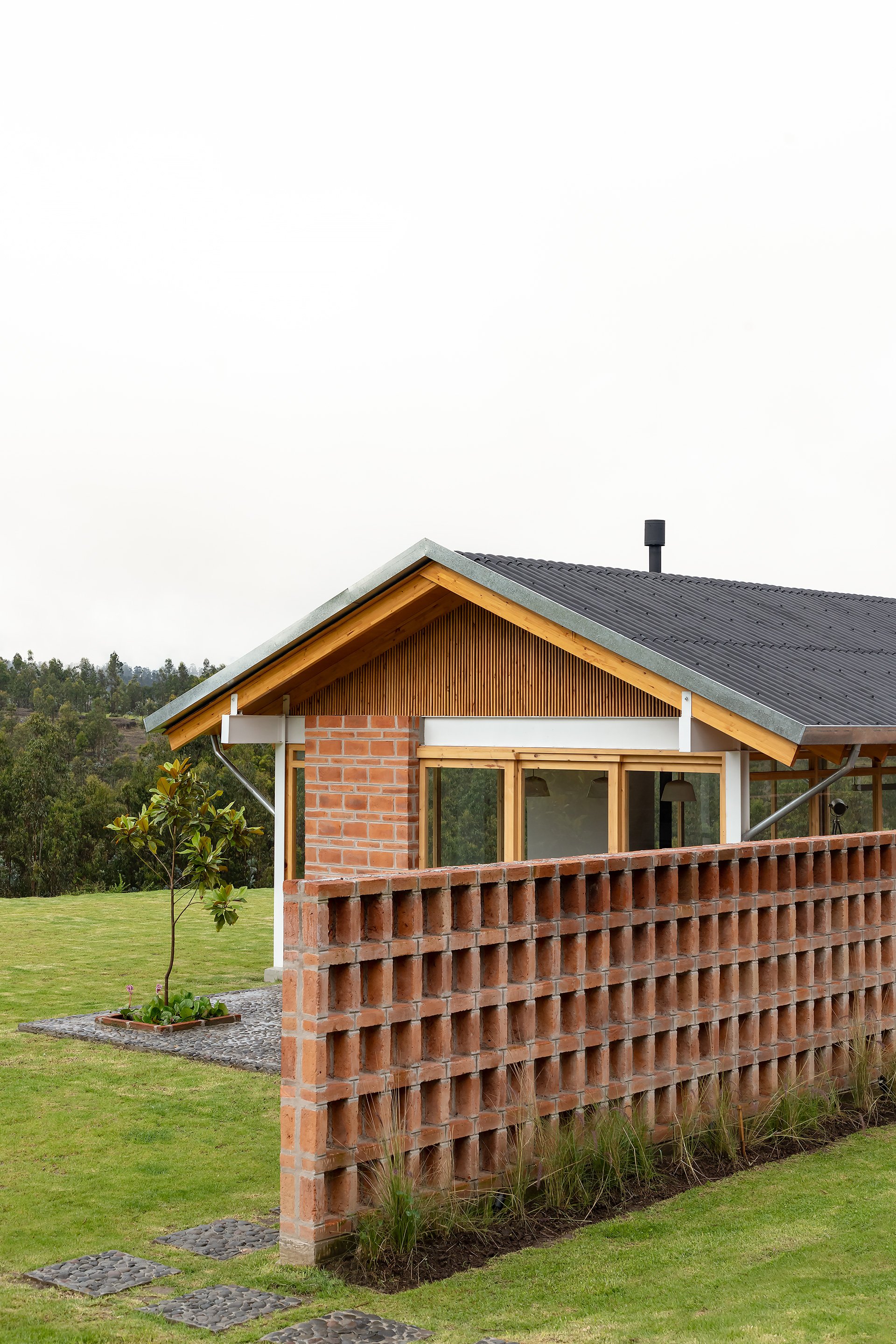 Casa moderna de madera con muro de ladrillos en un entorno natural con árboles y césped.