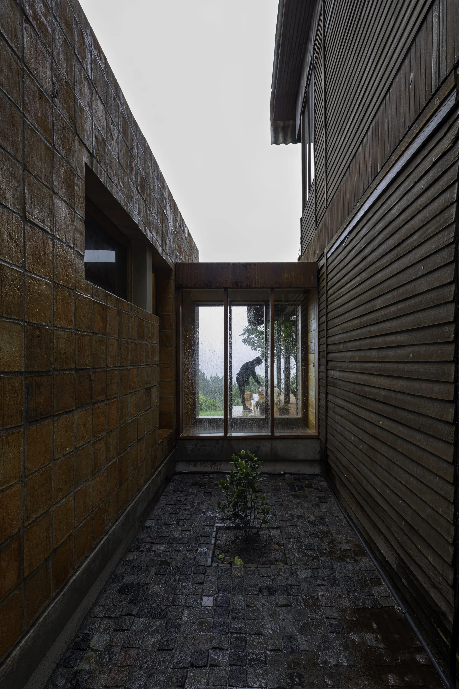 Paredes de ladrillo y madera en un pasillo abierto con piso de piedra, con plantas y una estructura de vidrio y madera en el fondo, y una persona en el interior, visible a través del vidrio, en un día lluvioso.