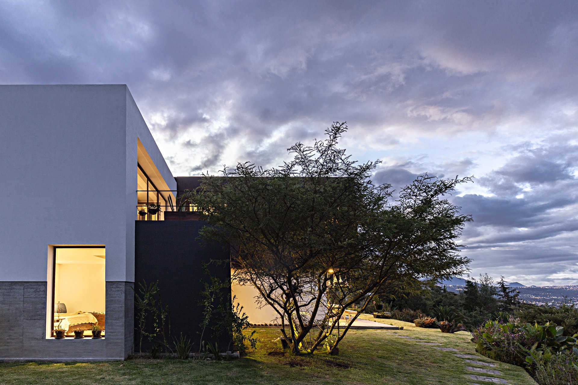 Casa moderna con luces encendidas, rodeada de árboles y jardín, con cielo nublado al atardecer