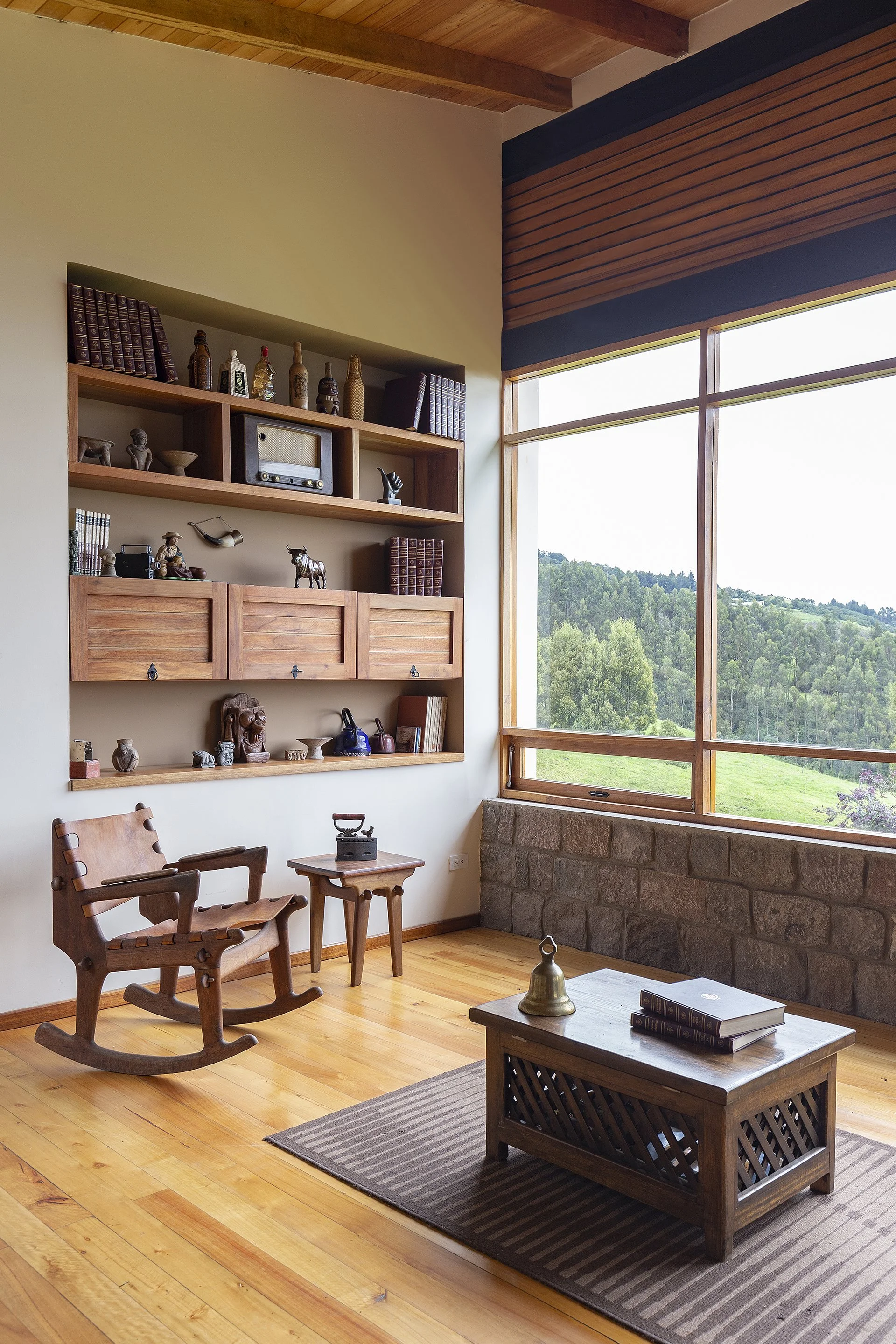 Sala con vista a la naturaleza, muebles de madera, estantería con libros y decoraciones, sillón de madera, mesa ratona con libros y objeto de metal, ventana grande con vista a árboles y colinas.