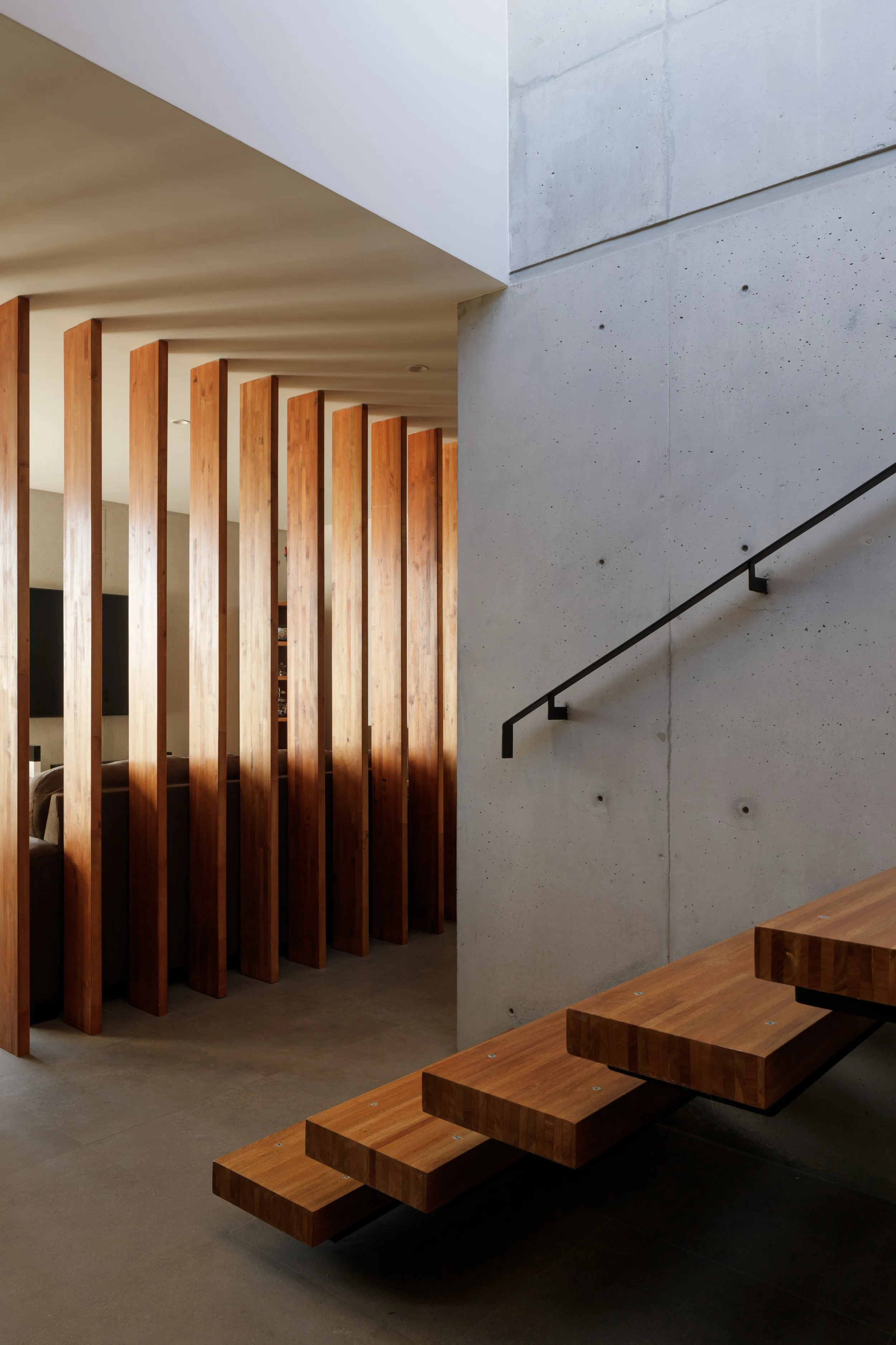 Escalera de madera con escalones flotantes en un espacio interior moderno, con pared de concreto y divisores de madera verticales.