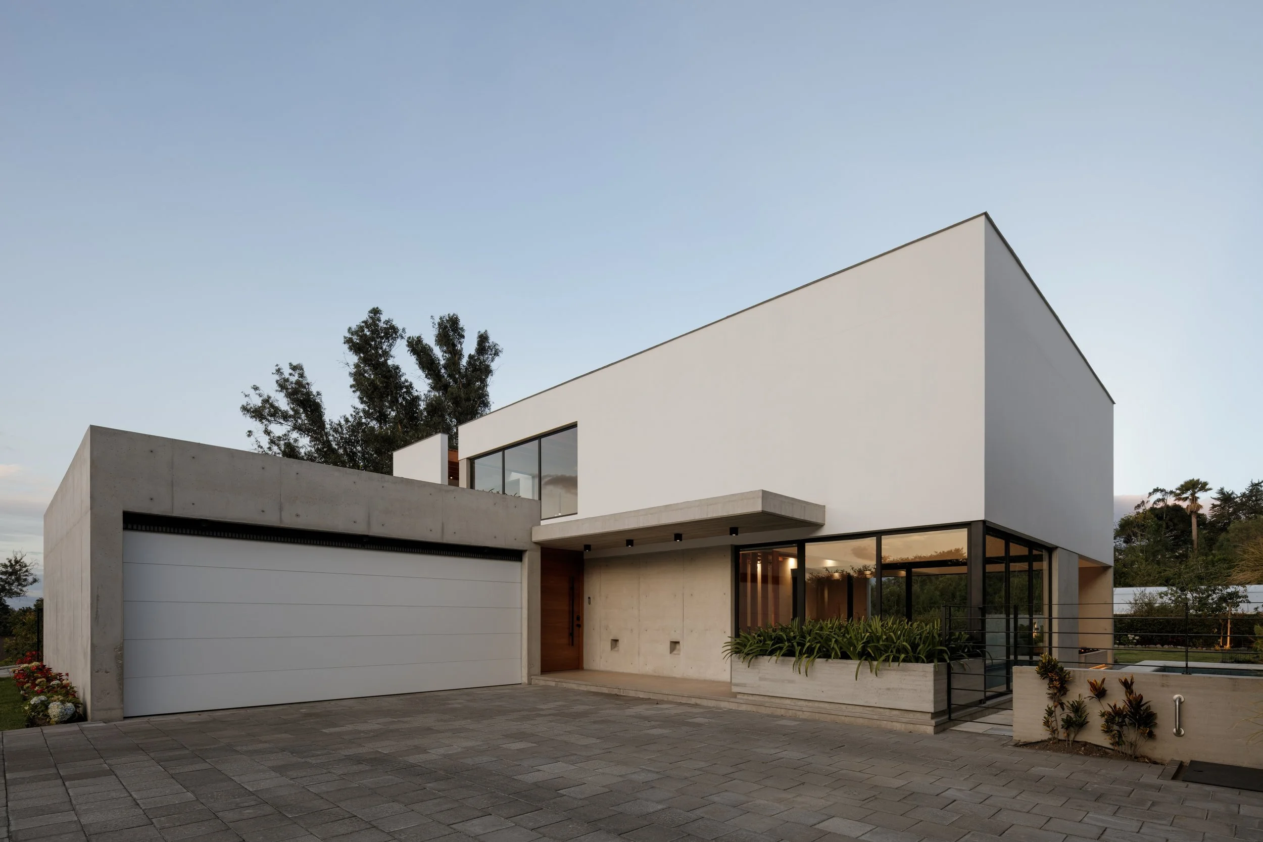 Casa moderna de dos pisos con estructura de concreto y muchas ventanas, jardín y garaje, con árboles y cielo despejado en el fondo.