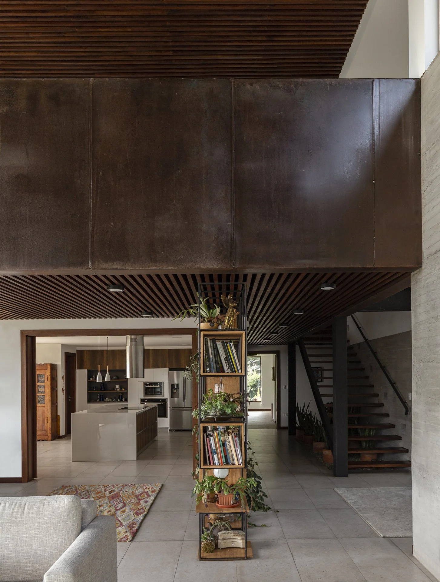 Sala moderna con estantería de plantas y libros, cocina en el fondo, paredes de piedra y madera en el techo y escalera de madera y metal a la derecha.