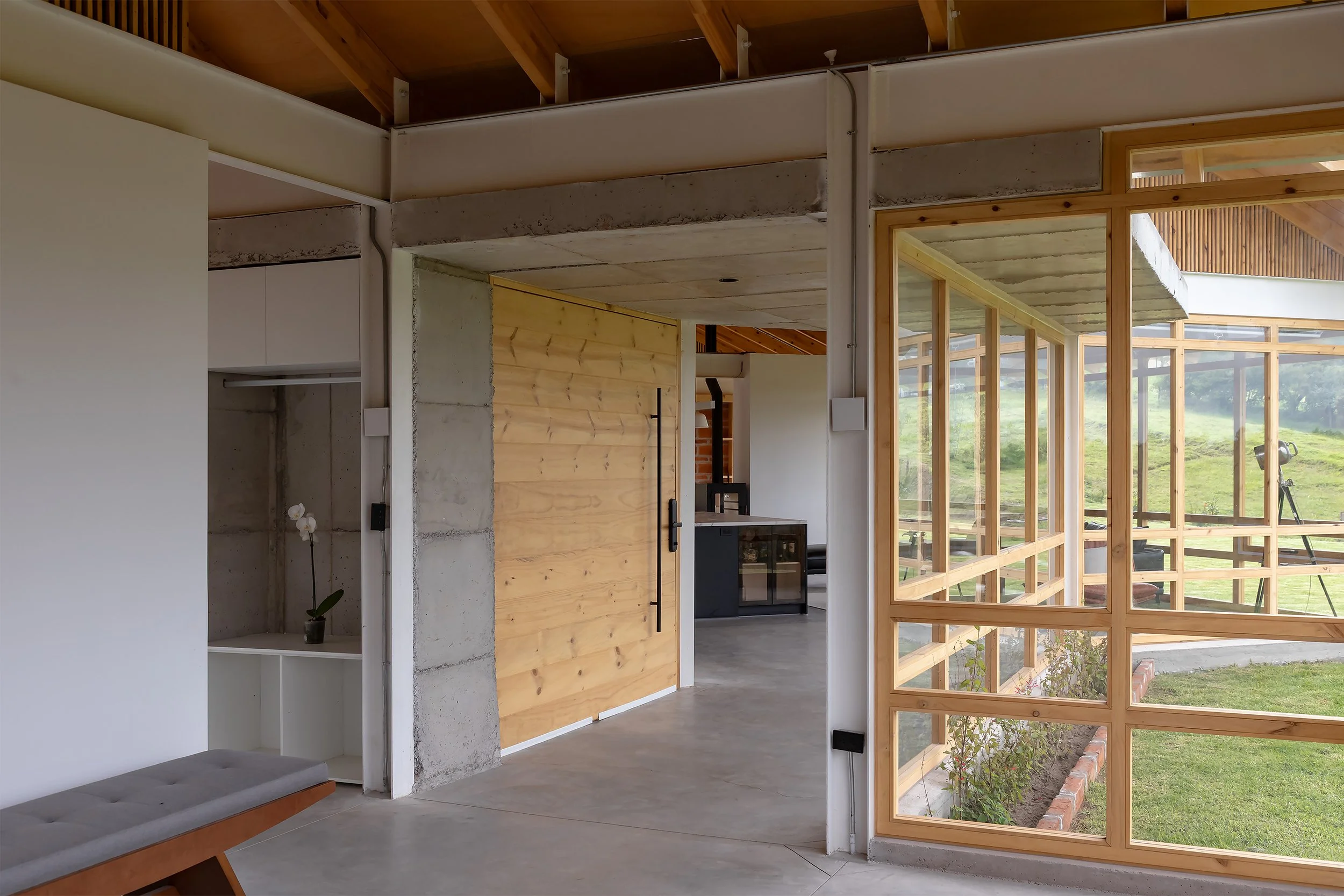 Interiores modernos con paredes de cemento y madera, grandes ventanas de madera que muestran un paisaje natural exterior, y un espacio de sala con muebles minimalistas.