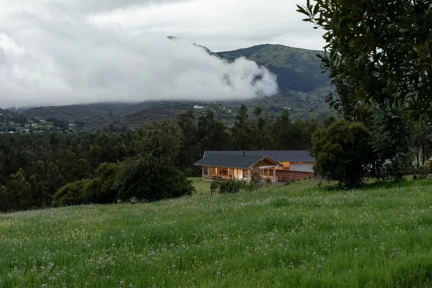 Casa de madera en un paisaje verde con árboles y montañas con niebla en el fondo bajo un cielo nublado.