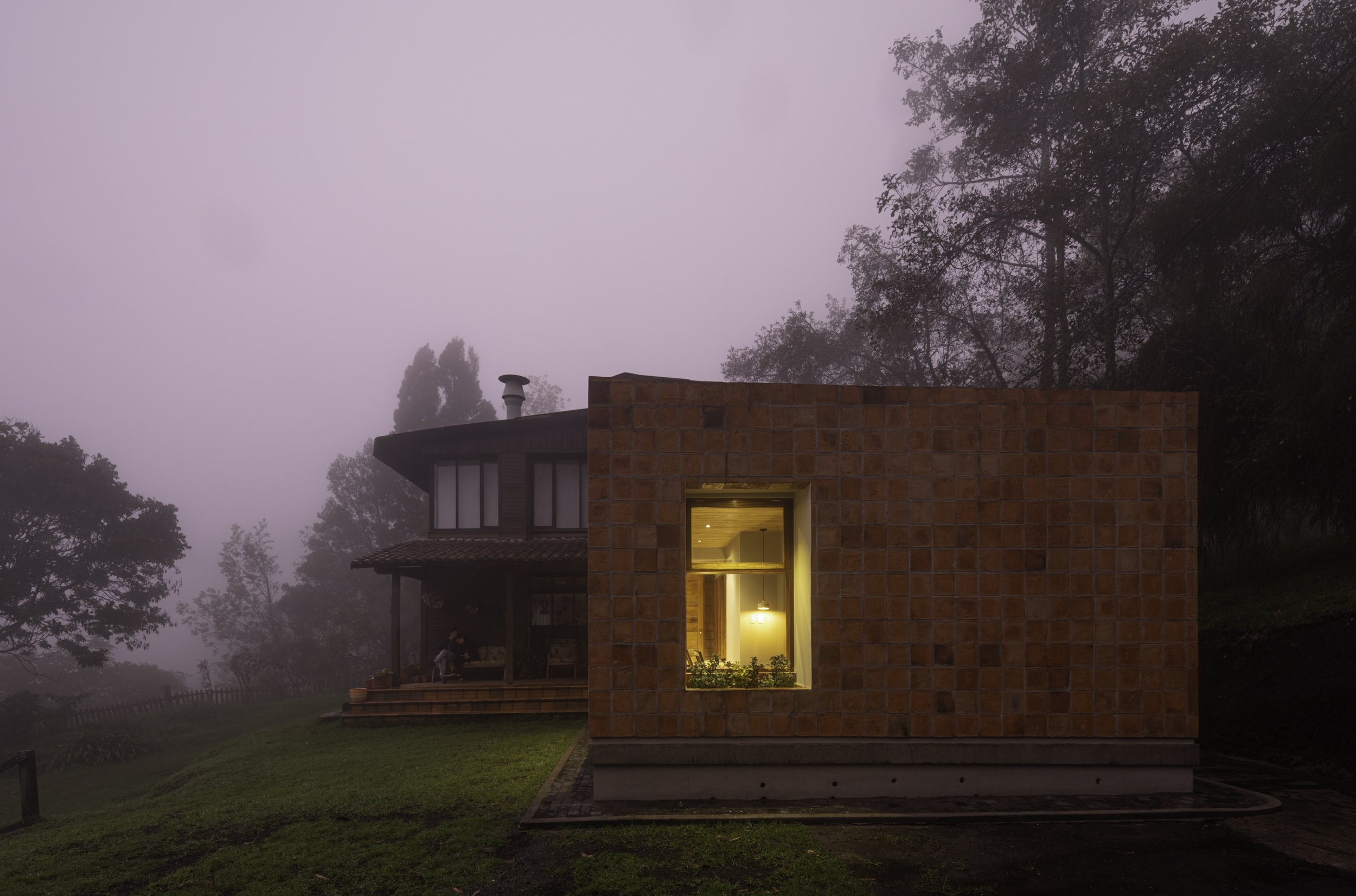 Casa moderna de ladrillo con ventana iluminada, rodeada de árboles y niebla en el fondo.