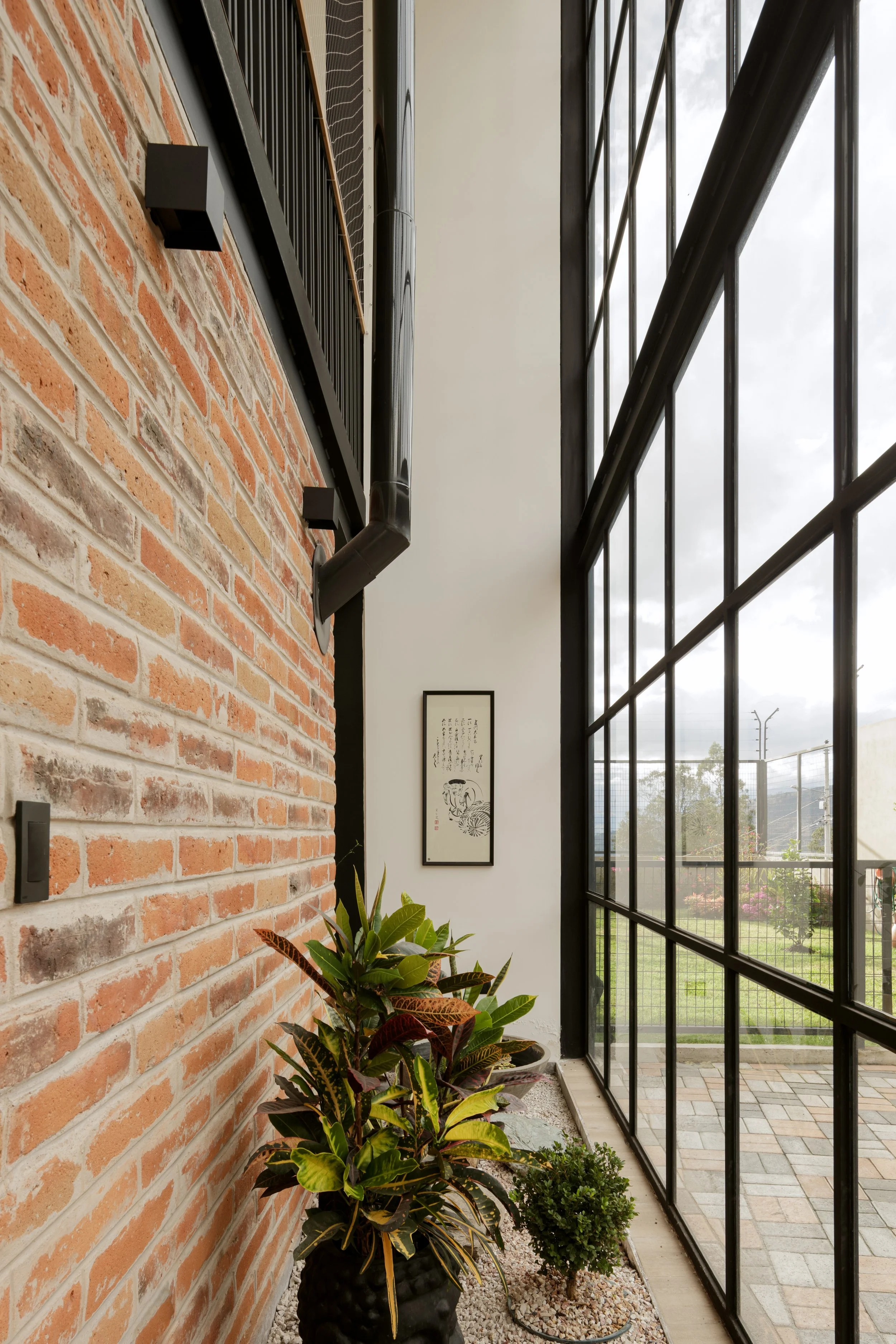 Un pasillo interior con una pared de ladrillo a la izquierda, plantas en macetas negras y una ventana grande de cristal con estructura de metal negro a la derecha, vista exterior de un área verde con cercado y cielo nublado.