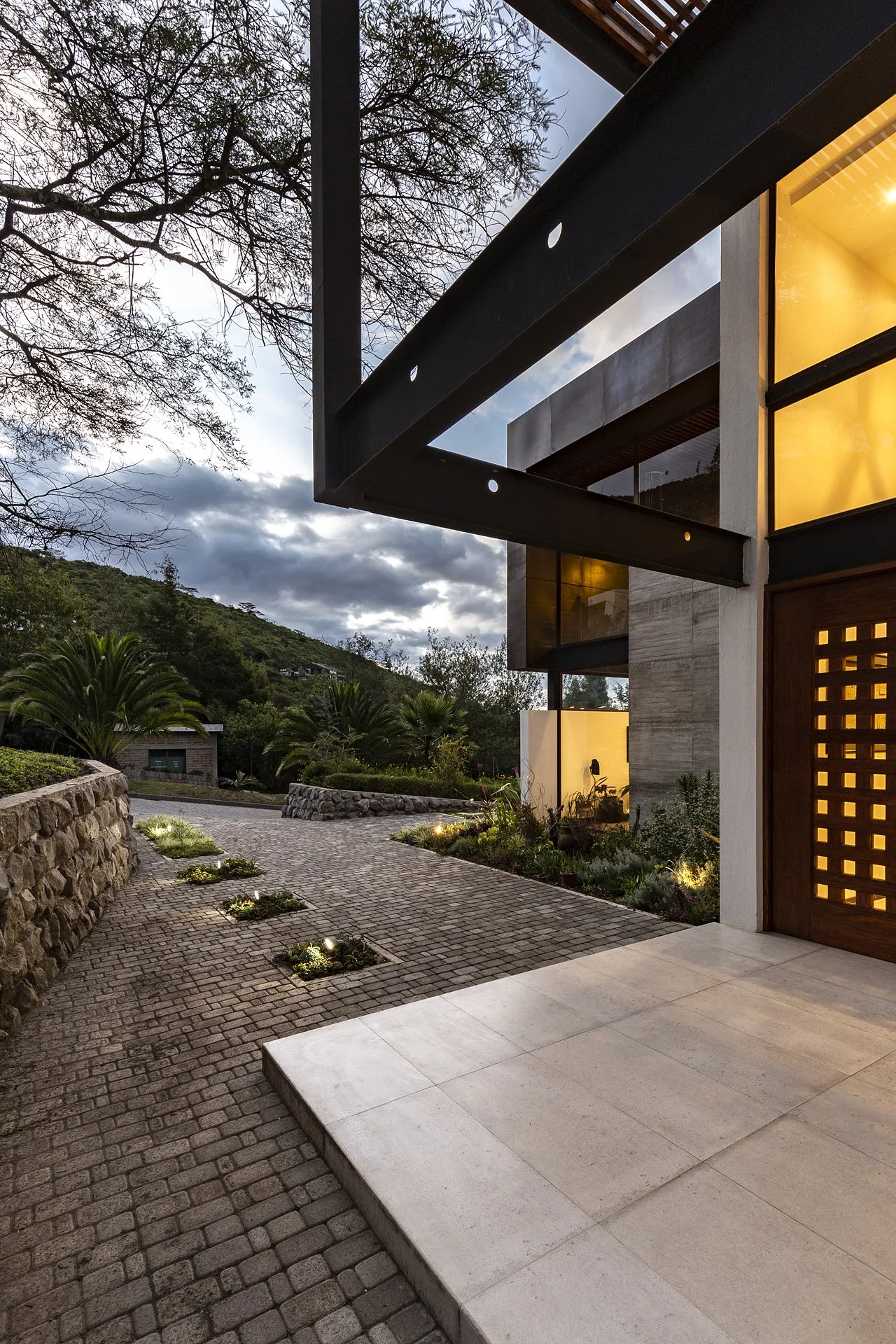 Vista del exterior de una casa moderna con iluminación nocturna, camino de piedra y jardín con plantas y árboles en un entorno montañoso.
