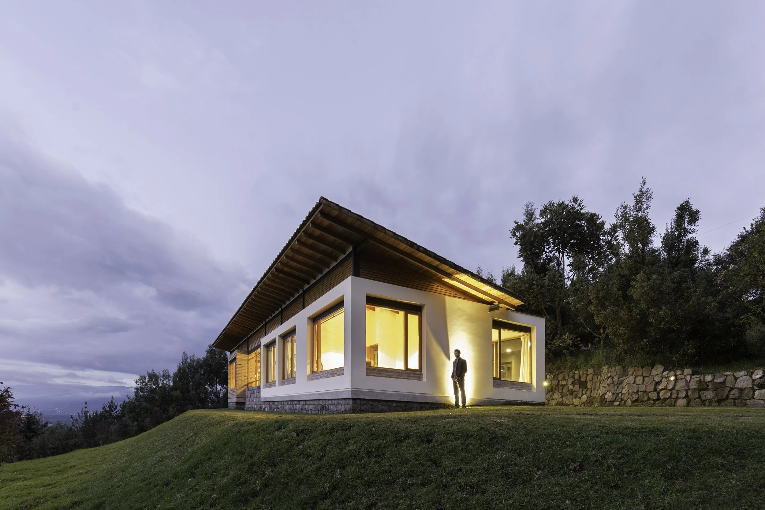 Casa moderna de dos pisos con ventanas grandes, iluminada desde adentro, ubicada en un área con césped y árboles, en la tarde con cielo nublado.