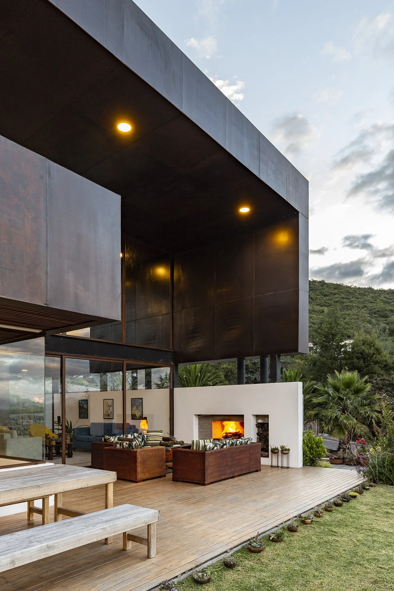 Exterior de una casa moderna con balcón de metal, chimenea encendida y muebles de patio de mimbre, rodeada de vegetación y jardines con cielo despejado y nubes.
