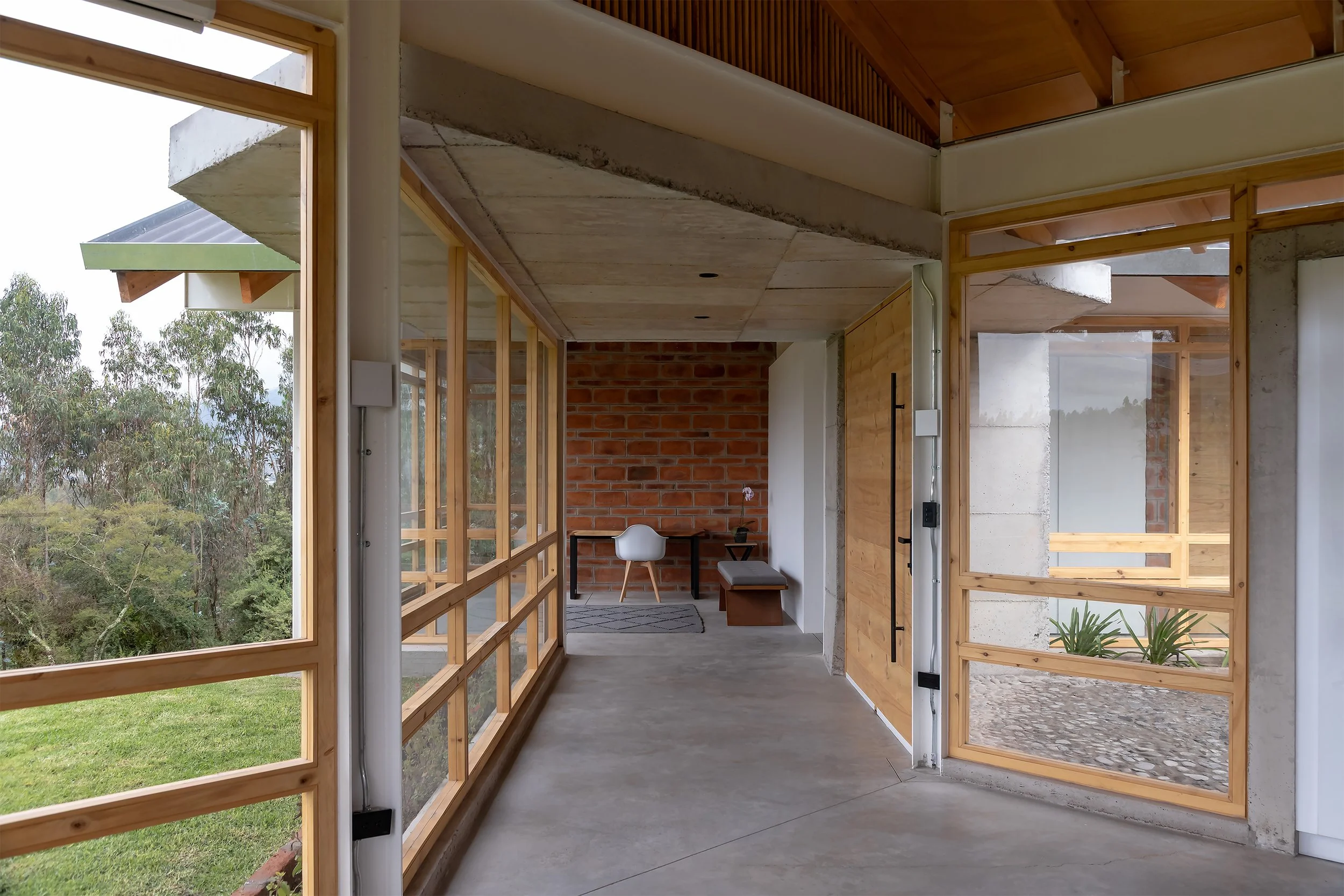 Interior de una casa en construcción con paredes de ladrillo y estructura de madera, ventanas de vidrio y vista a la naturaleza exterior.