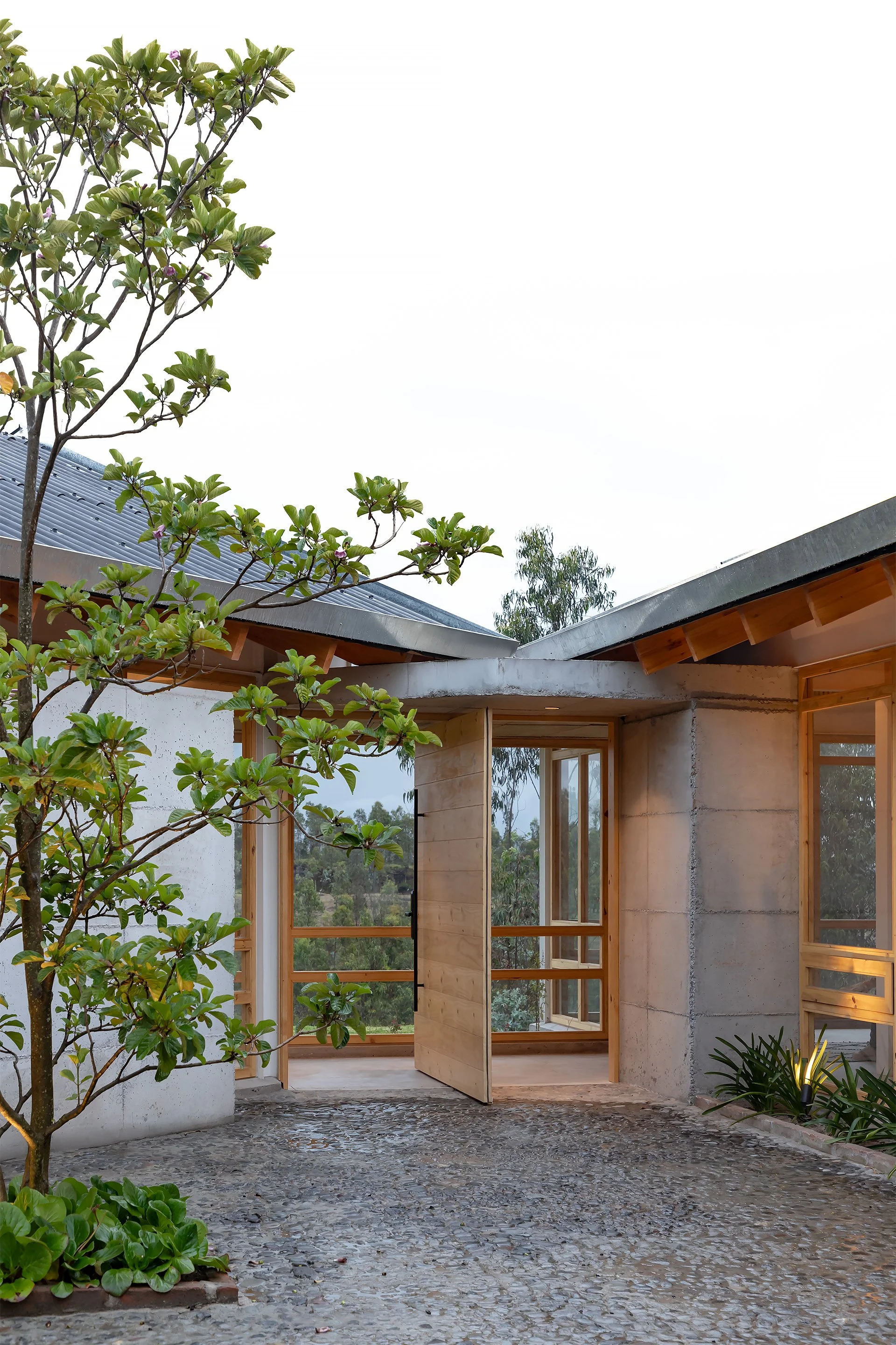 Vista de una casa moderna con ventanas de madera y un patio de piedra, rodeada de árboles verdes y plantas decorativas, con un cielo nublado.