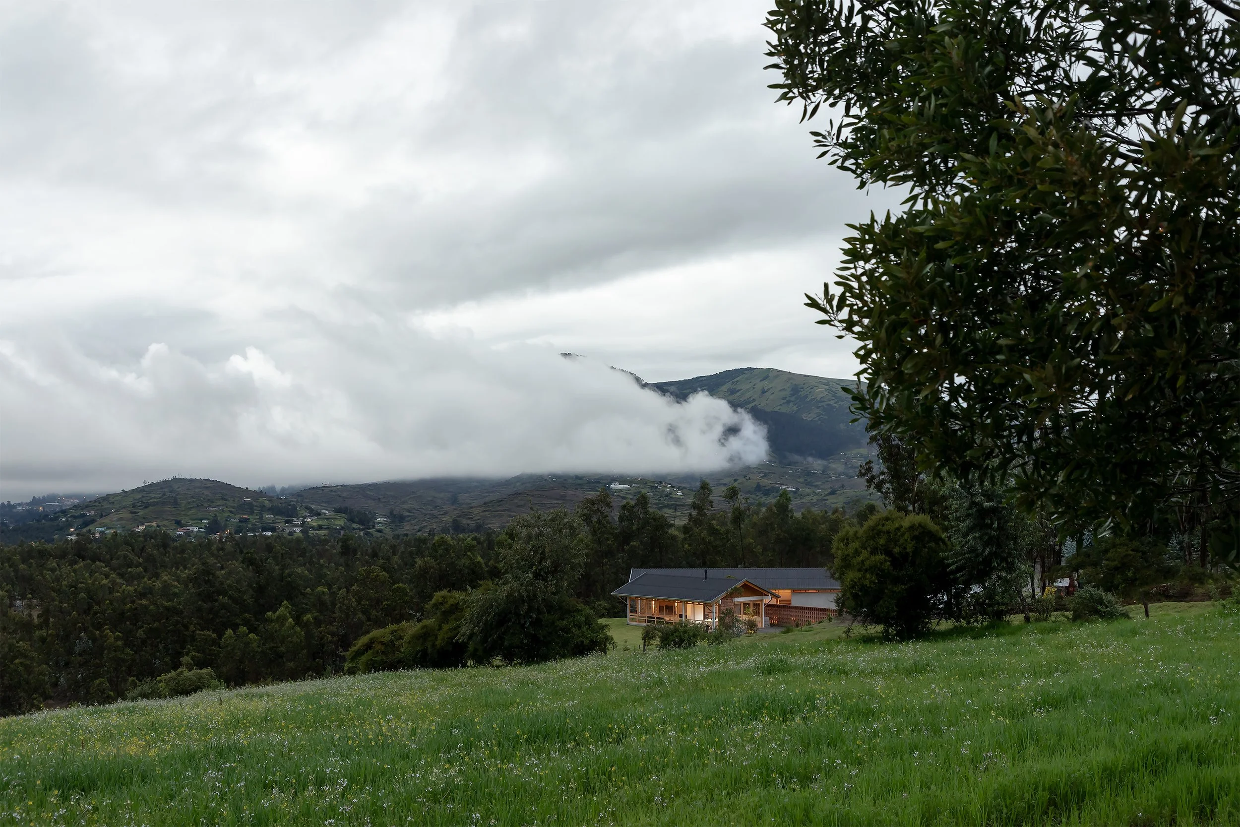 Paisaje montañoso con nubes bajas, un árbol grande a la derecha y una casa de madera en primer plano, en un entorno rural.