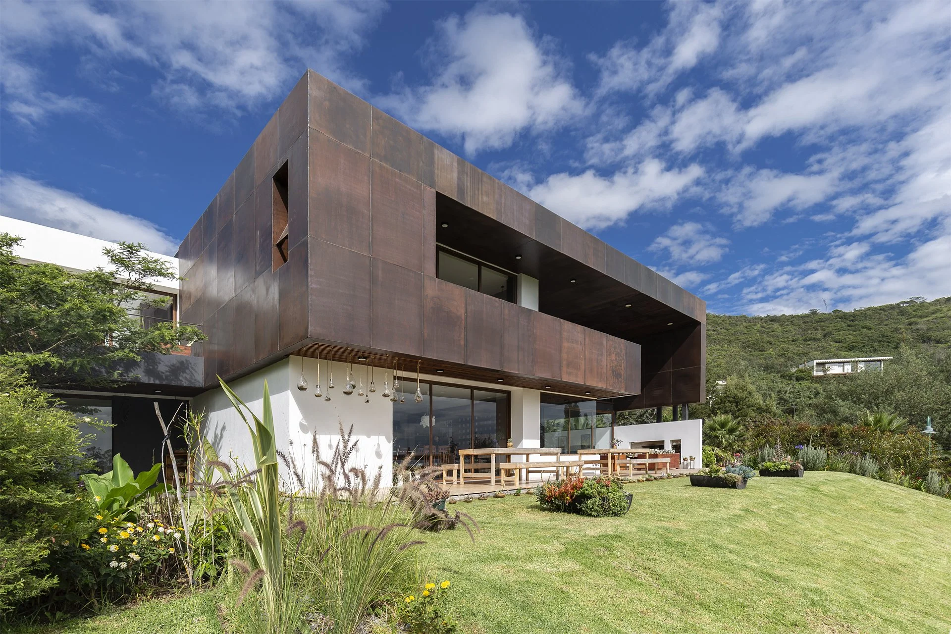 Casa moderna de dos pisos con paredes de metal, gran ventanal y balcón en un entorno natural con árboles y jardín bajo un cielo con algunas nubes.