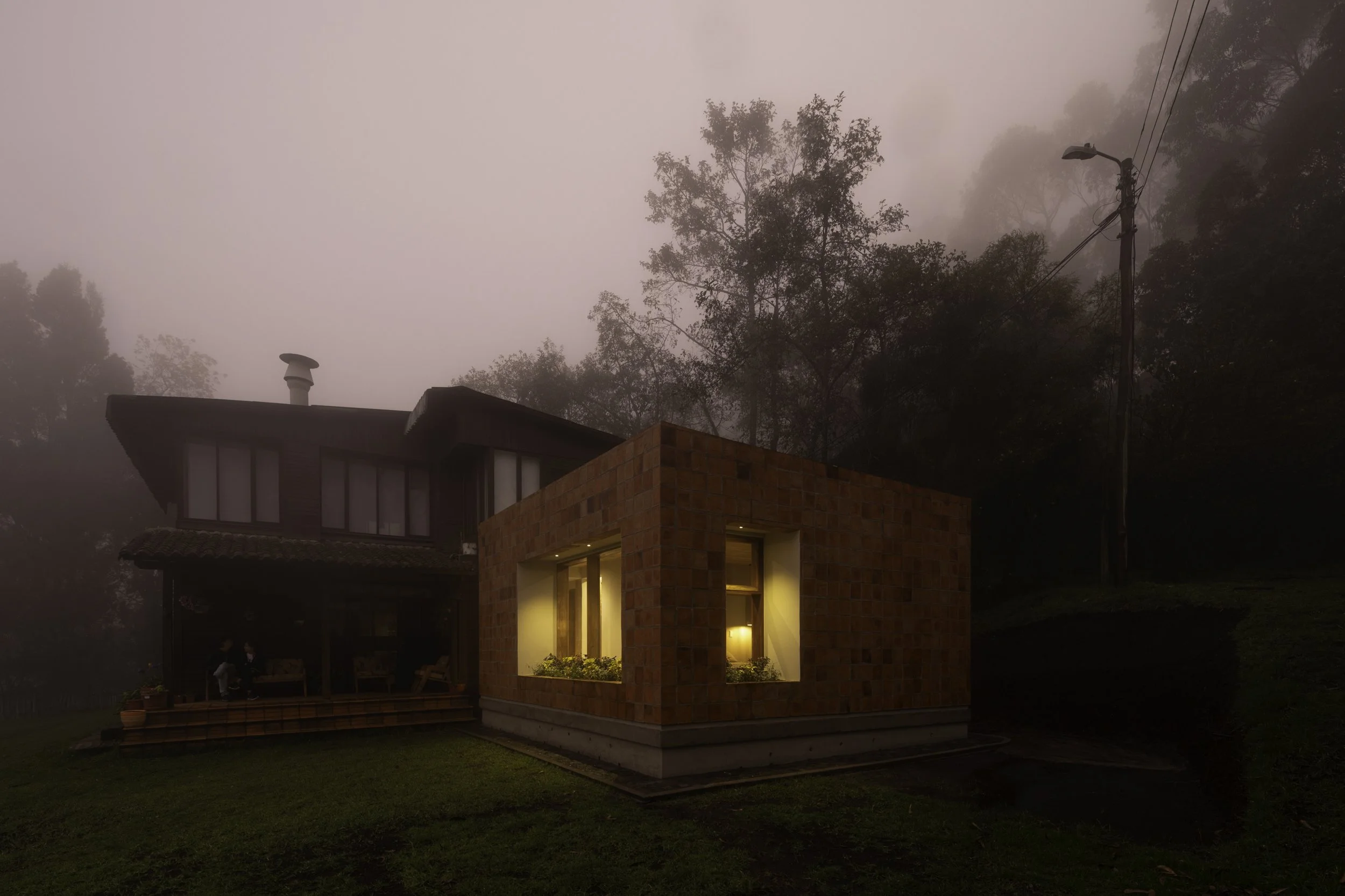 Casa moderna de dos pisos hecha con ladrillos, con ventanas iluminadas en un entorno de niebla y árboles en el fondo.