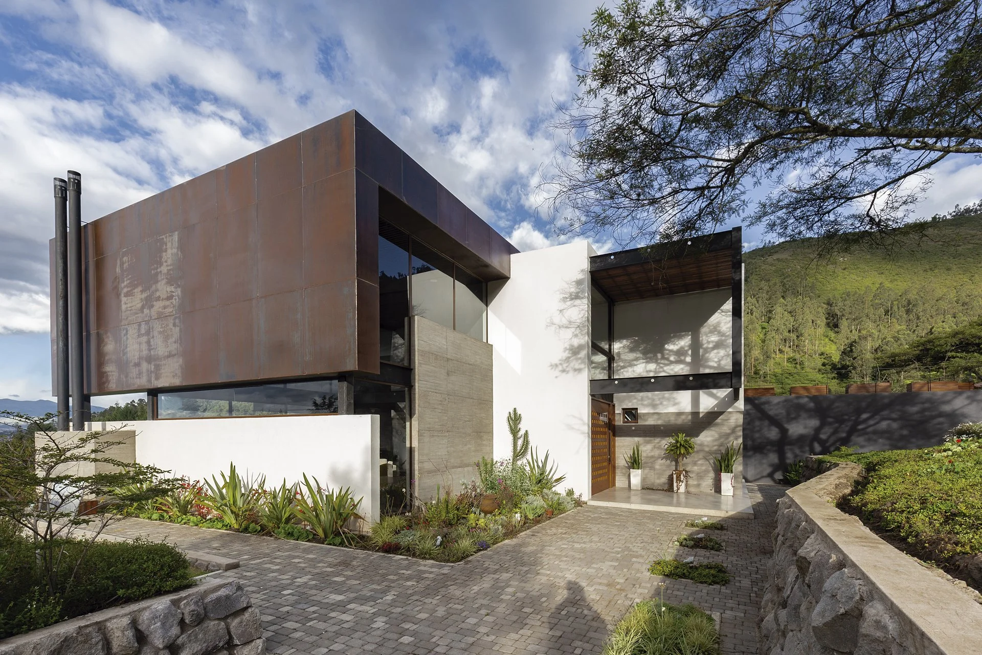 Casa moderna en un entorno natural con montañas y árboles, con un diseño arquitectónico contemporáneo de líneas rectas y materiales diversos.