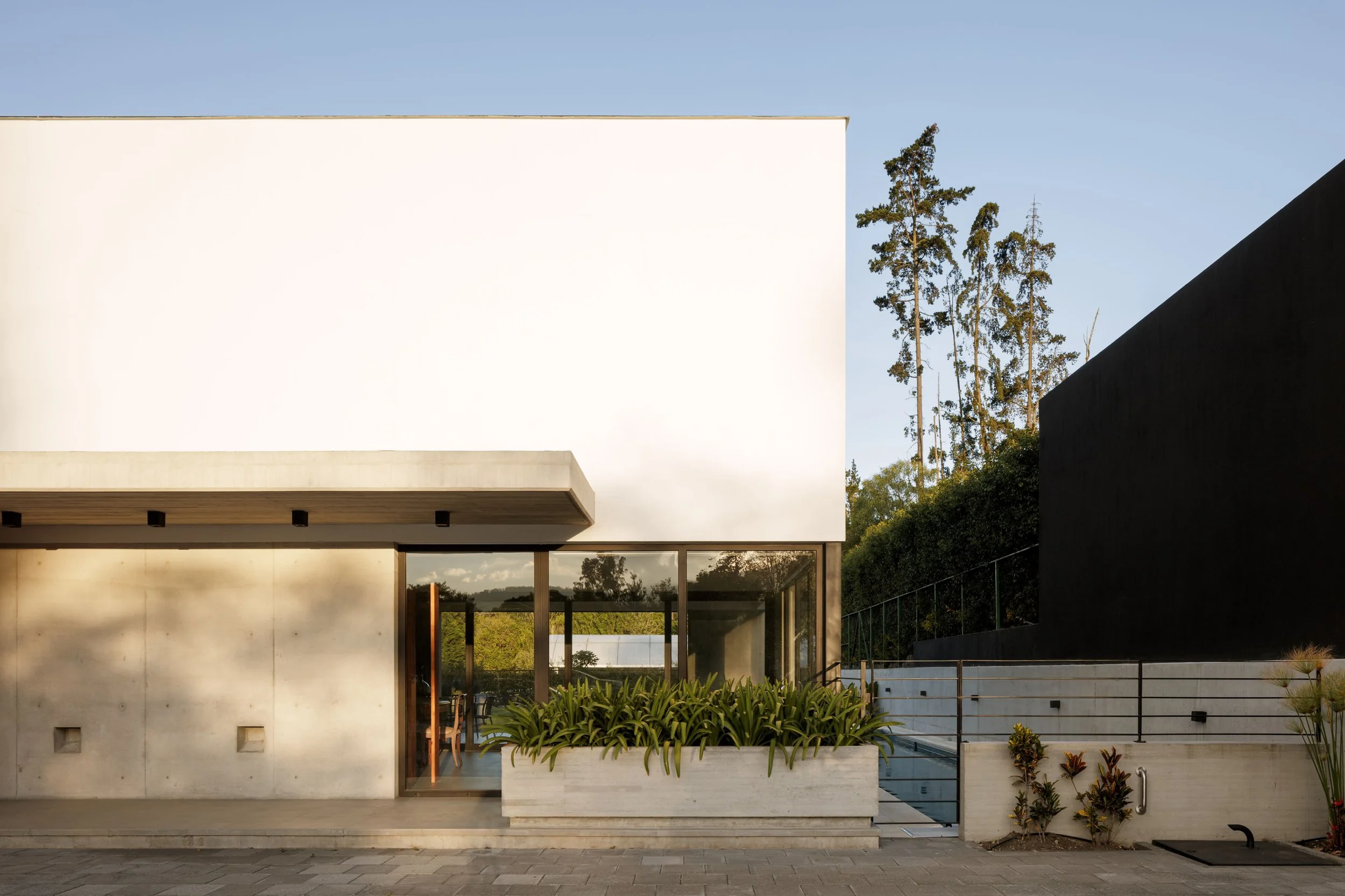 Vista frontal de una casa moderna con paredes blancas, ventanas de vidrio, un espacio para comer con sillas de madera y un jardín con plantas en la parte delantera