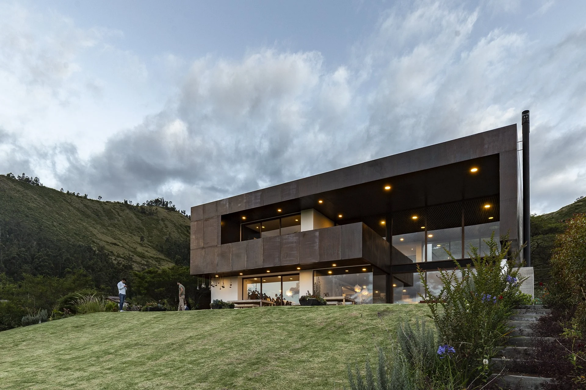 Vista de una casa moderna en un paisaje montañoso con cielo nublado y gente en el jardín.