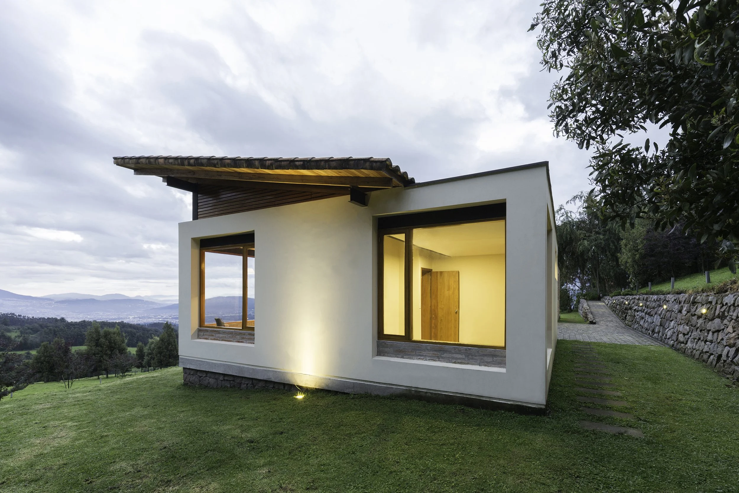 Casa moderna con ventanas grandes, iluminada desde el interior, en un entorno natural con césped, árboles y montañas al fondo, cielo nublado.
