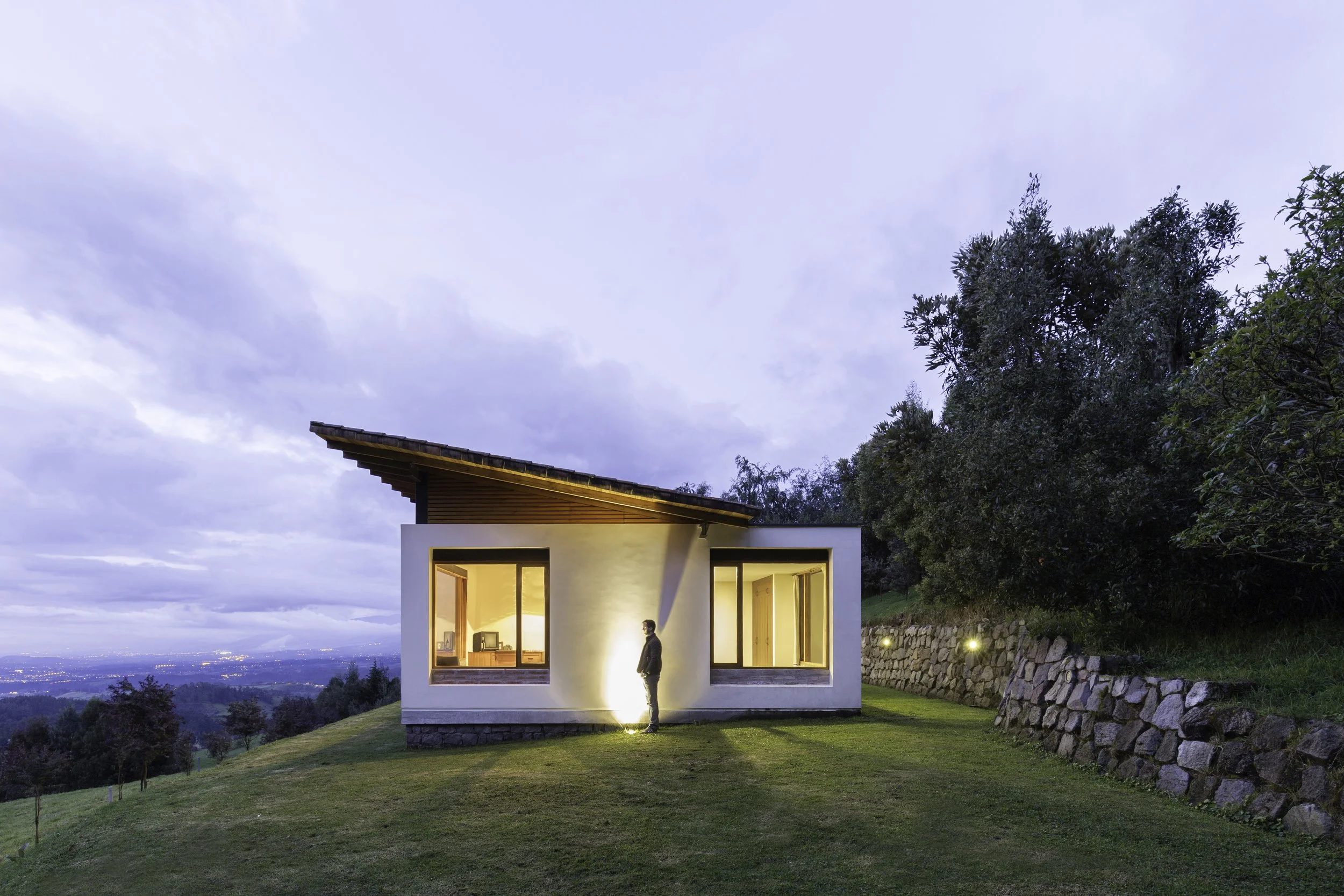 Casa moderna con ventanas iluminadas en una colina, rodeada de árboles y césped, con cielo nublado al atardecer.