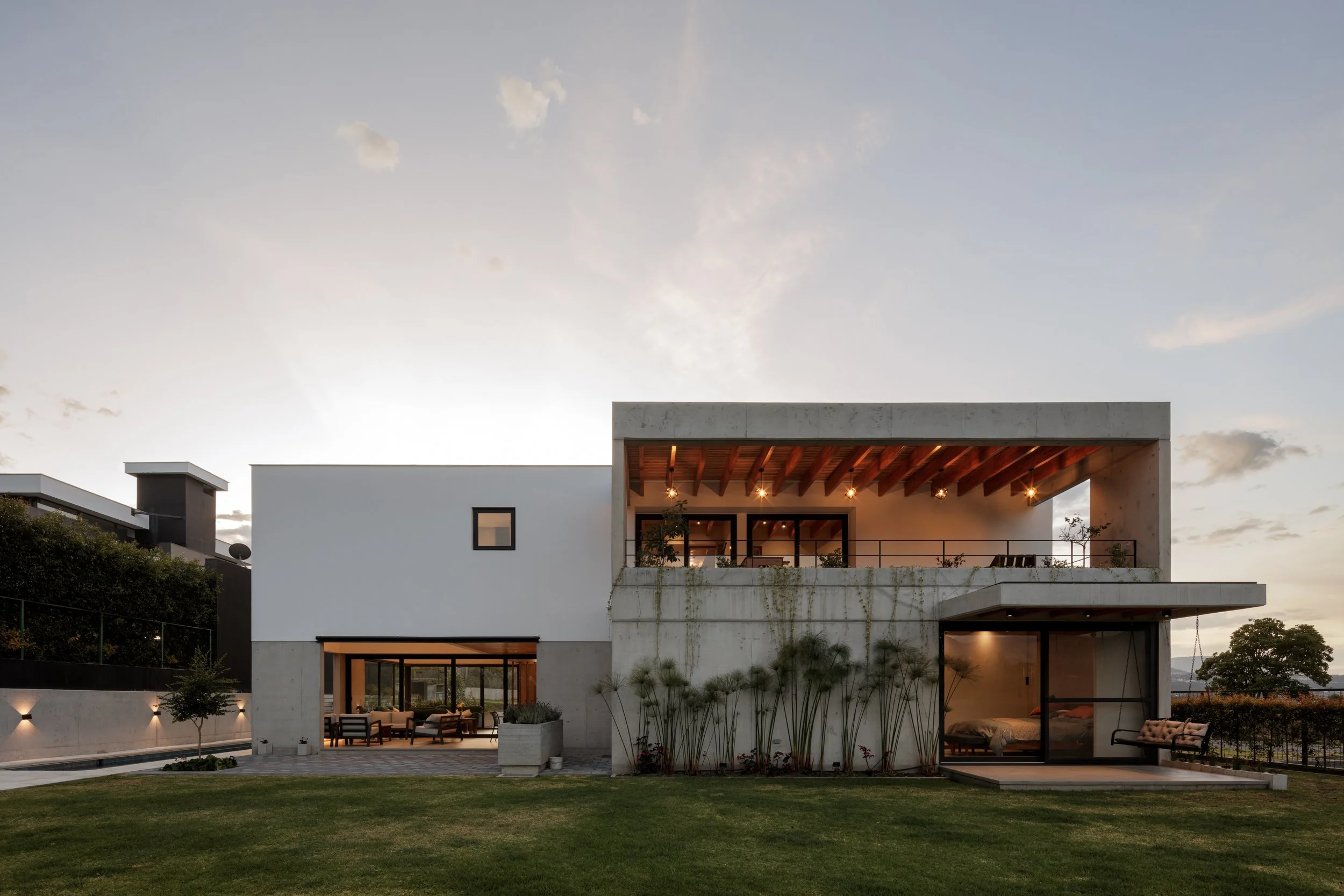 Casa moderna de dos pisos con balcón, techos de madera y grandes ventanales, rodeada de jardín con césped y plantas, al atardecer con cielo claro y algunas nubes.