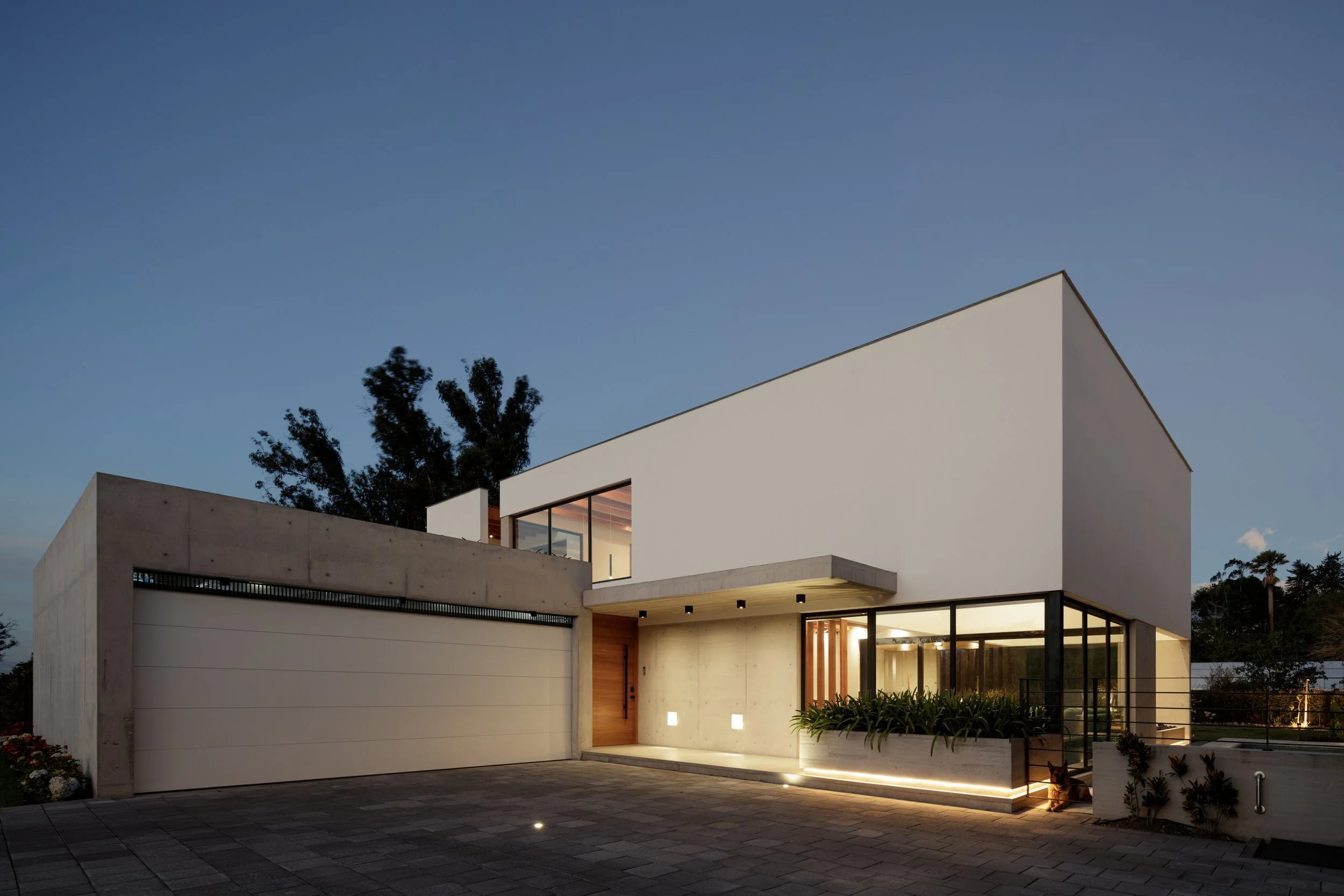 Fachada moderna de una casa de dos pisos con paredes blancas, ventanas grandes, iluminación exterior y un área de entrada con plantas ornamentales. Al fondo, árboles y cielo despejado al atardecer.