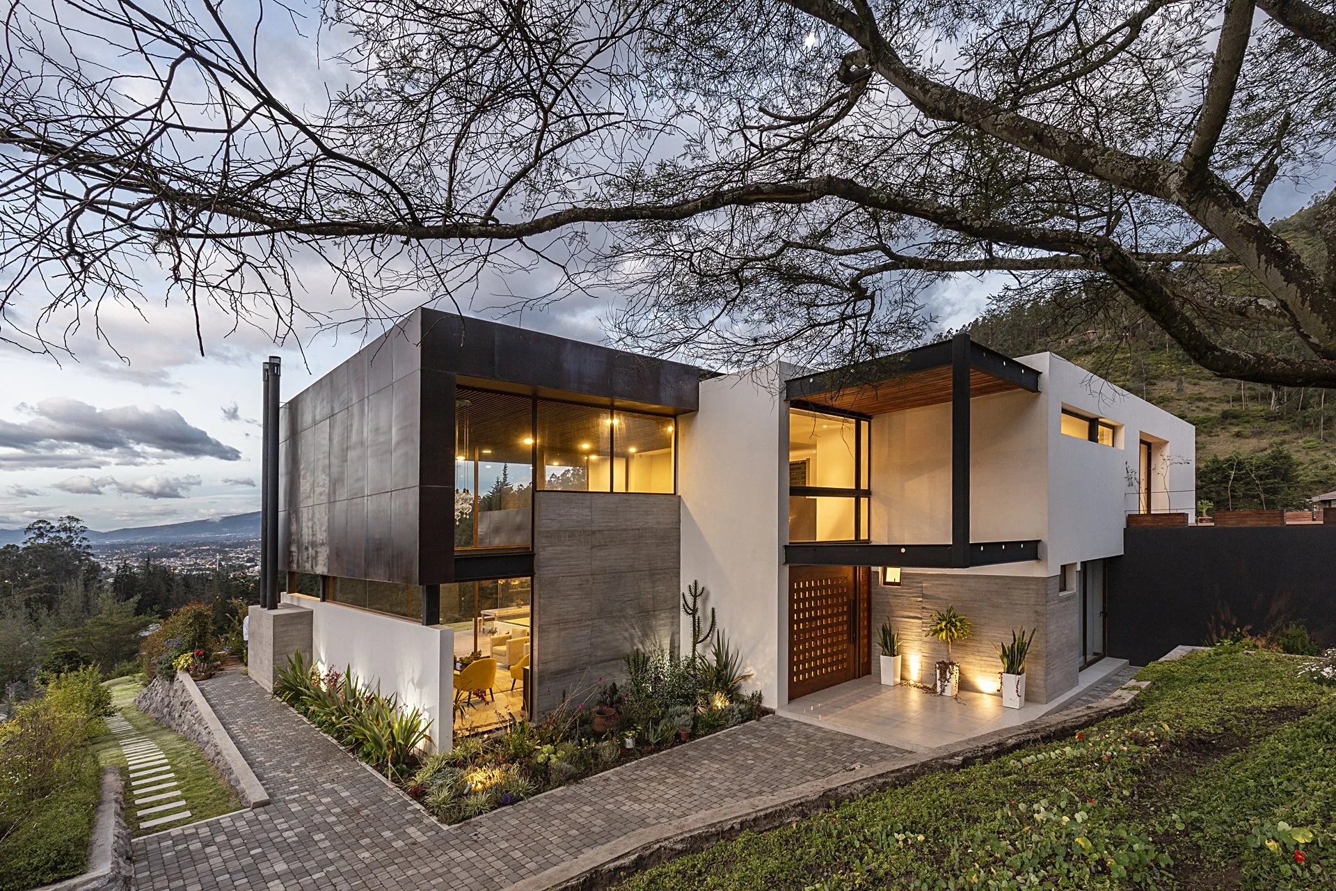 Casa moderna en un entorno natural con árboles y paisaje montañoso al atardecer, iluminación interior visible por ventanas grandes
