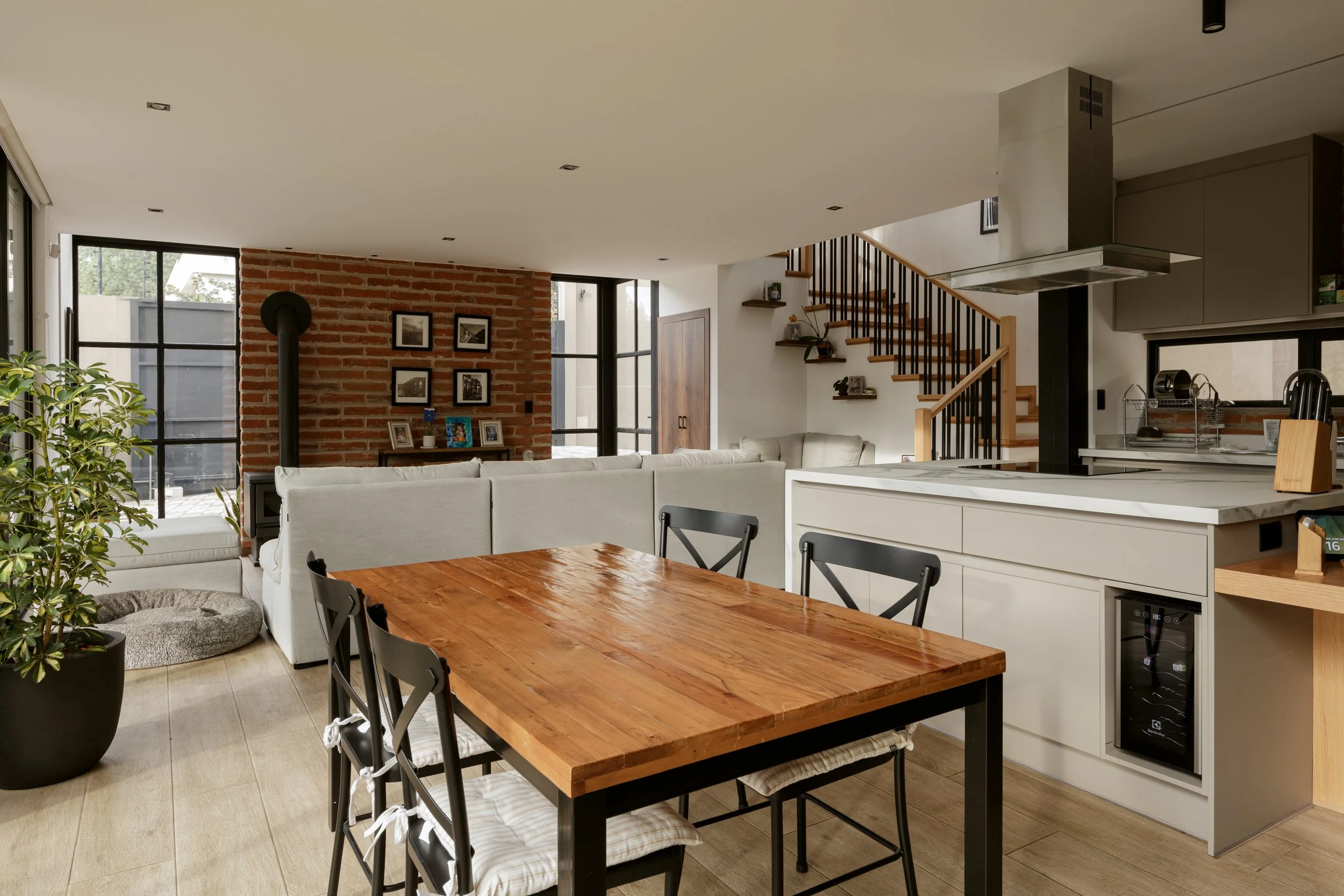 Interior de una casa moderna con sala y cocina, paredes de ladrillo, ventanas grandes, muebles blancos y madera, plantas y escaleras con barandilla de madera y metal.
