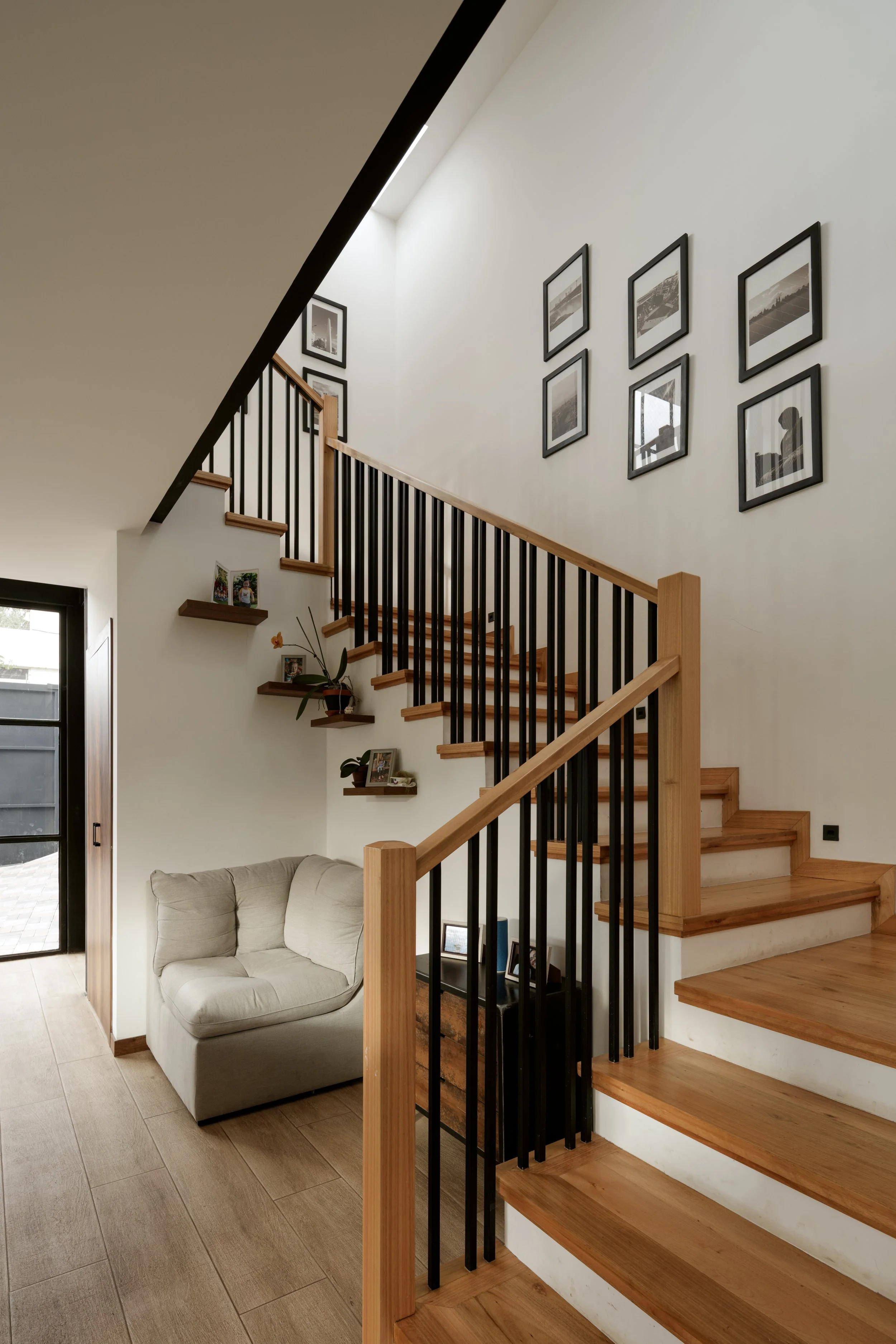 Interior de una sala con escalera de madera y barandilla negra, paredes blancas con cuadros decorativos y una esquina con estantes con fotos, planta y sofá beige junto a una puerta de vidrio.