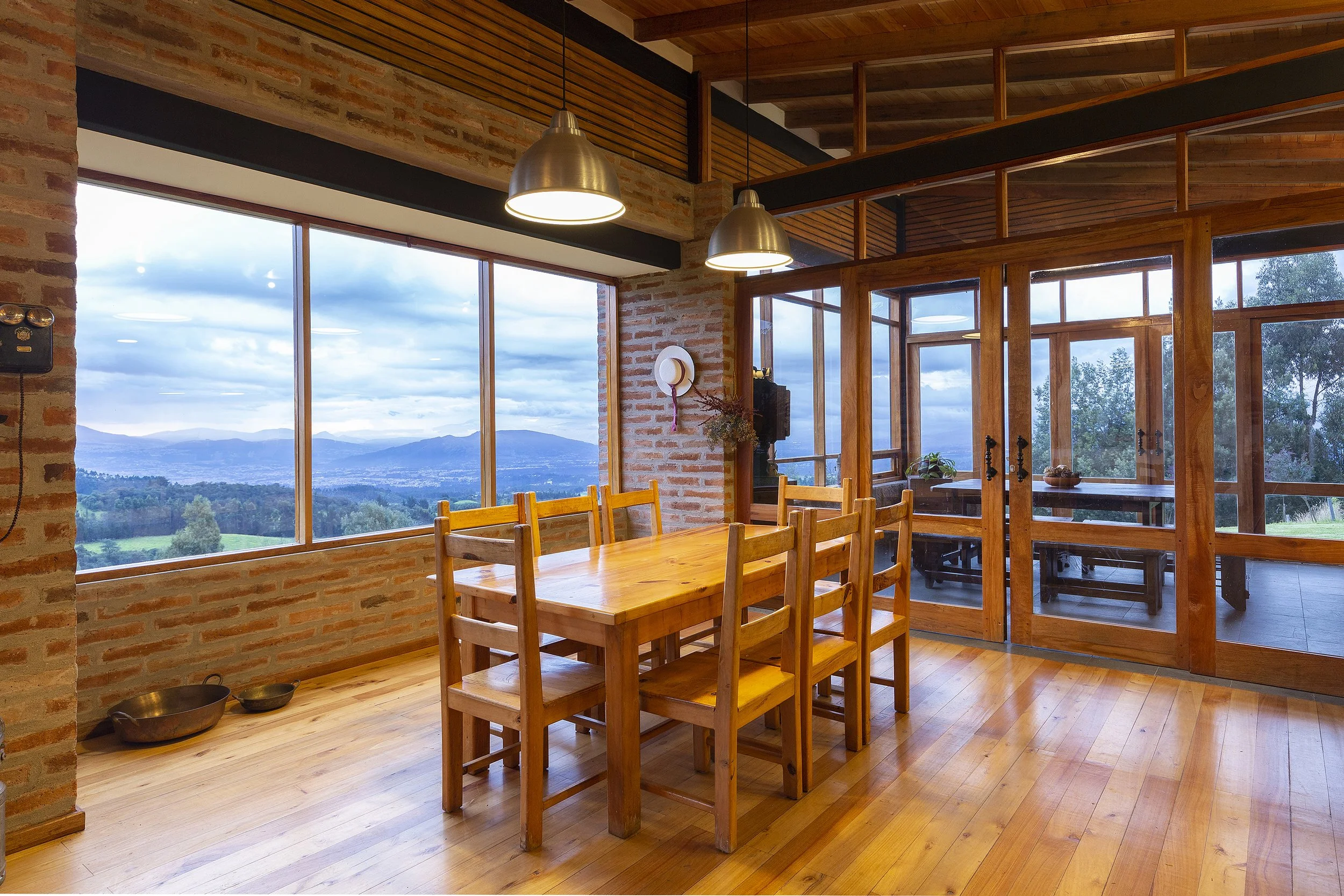 Interior de un comedor con mesas de madera, ventanas grandes con vista a montañas y árboles, paredes de ladrillo y estructura de madera, colgantes de metal y lámparas de techo con luz cálida.
