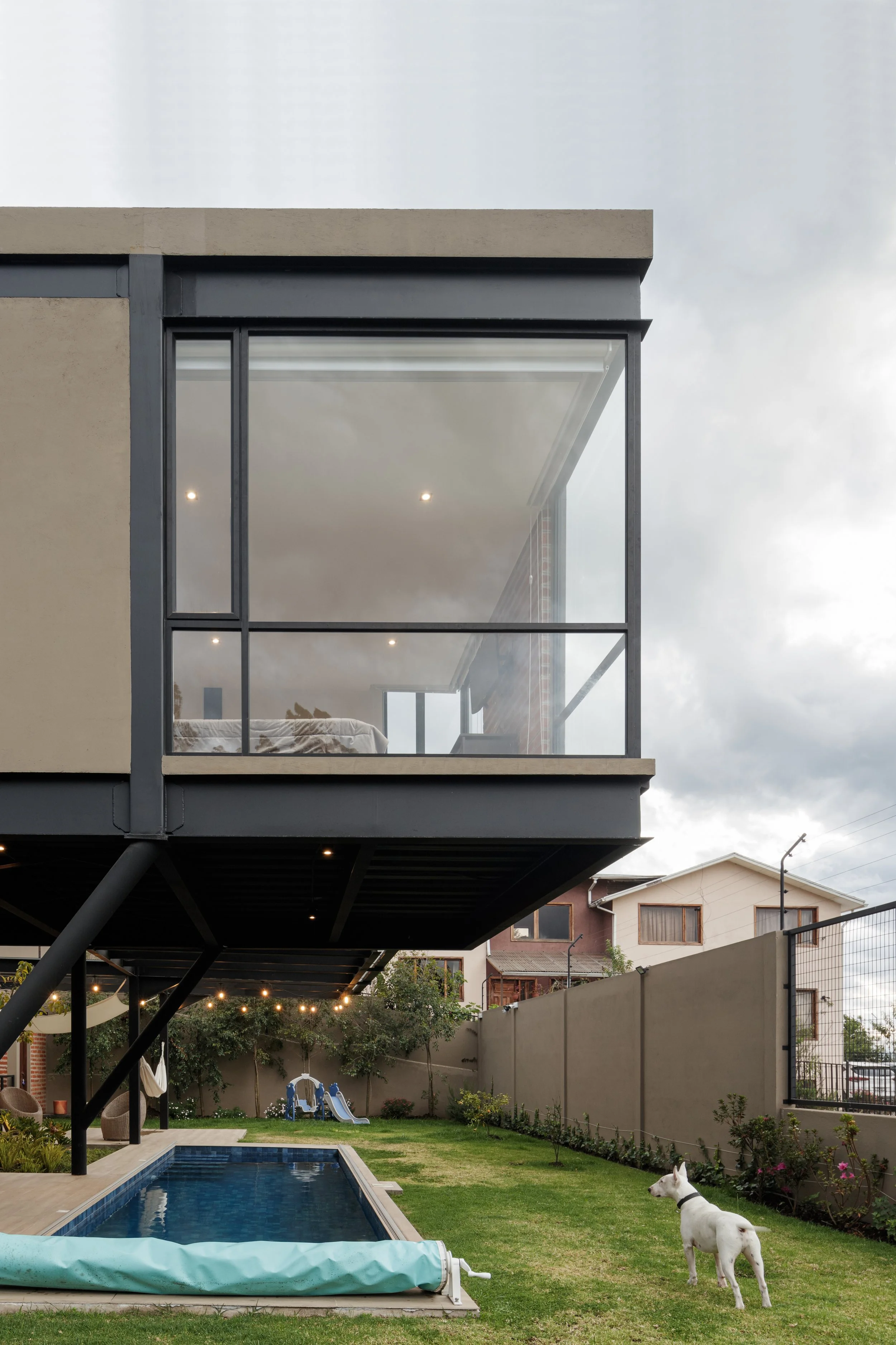 Casa moderna de dos niveles vista desde el patio trasero, con una piscina inflable cubierta y un perro blanco durmiendo cerca del césped, en un día nublado.