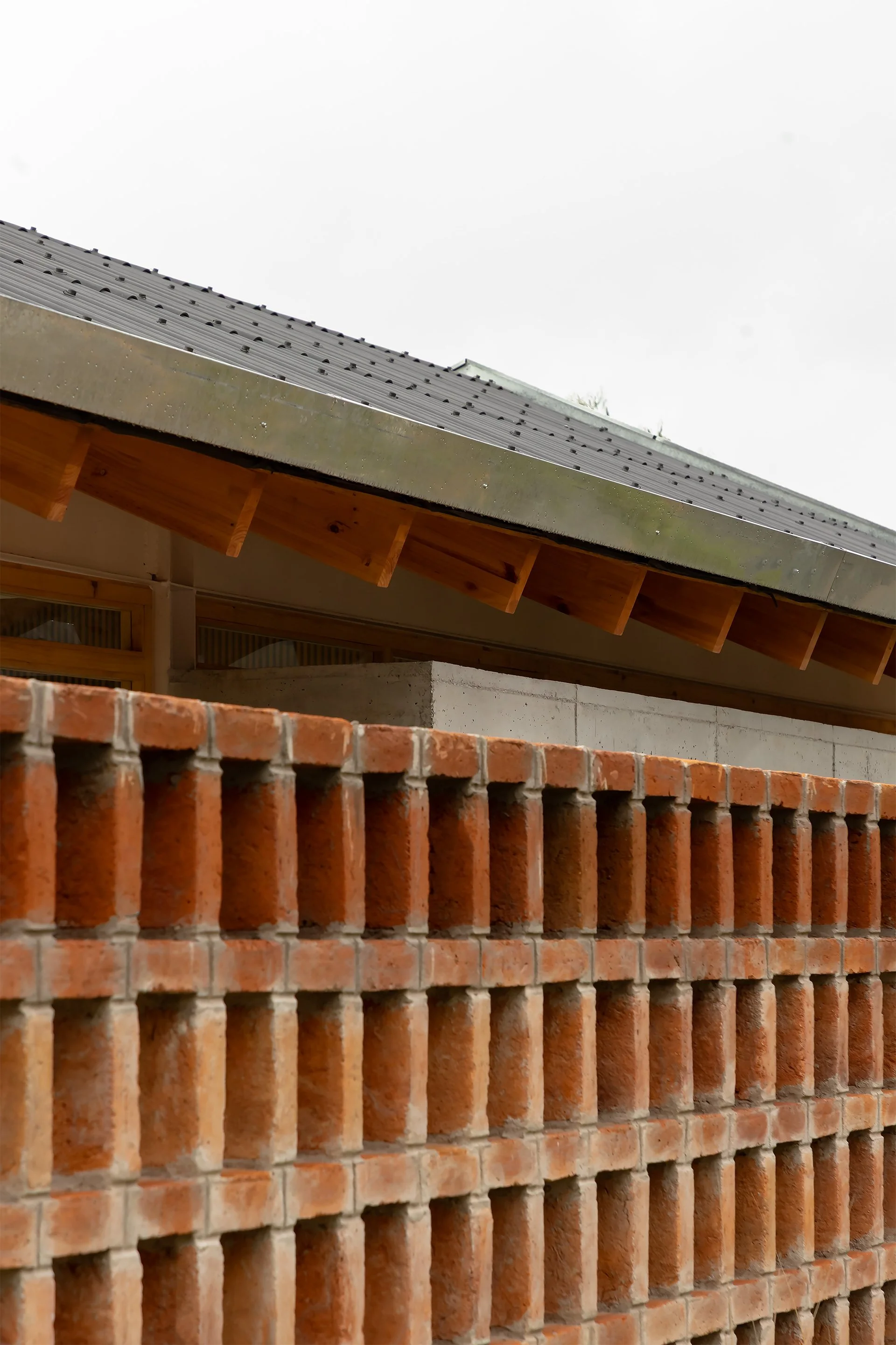 Vista de una obra en construcción con muro de ladrillos rojos en primer plano y estructura de madera y techo metálico en el fondo.